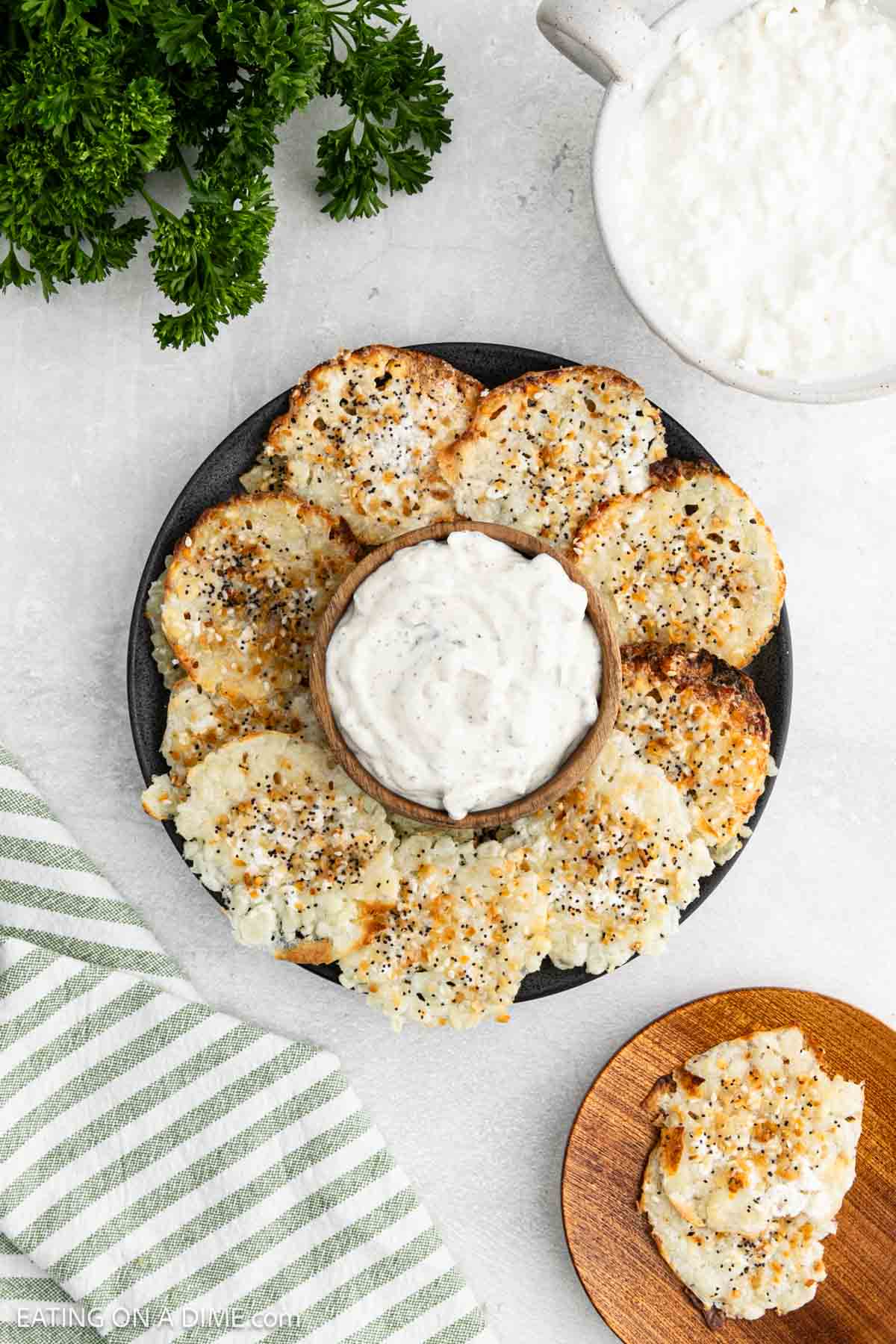 A round black plate with crispy cottage cheese chips arranged in a circle around a bowl of creamy dip. A bunch of parsley, a bowl of dip, and a striped towel are nearby on a light surface.