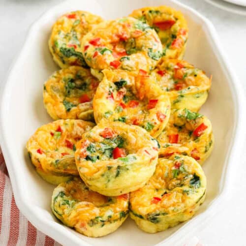 A white plate holds a stack of colorful Cottage Cheese Egg Bites filled with spinach, red bell peppers, and cheese, arranged neatly and ready to serve.