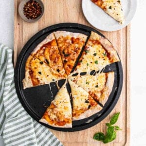 A pizza with melted cheese and herbs, made on a high protein pizza crust, is sliced into eight pieces on a black pan. One slice is on a small white plate. Basil leaves, red pepper flakes, and a striped towel are nearby on a wooden surface.