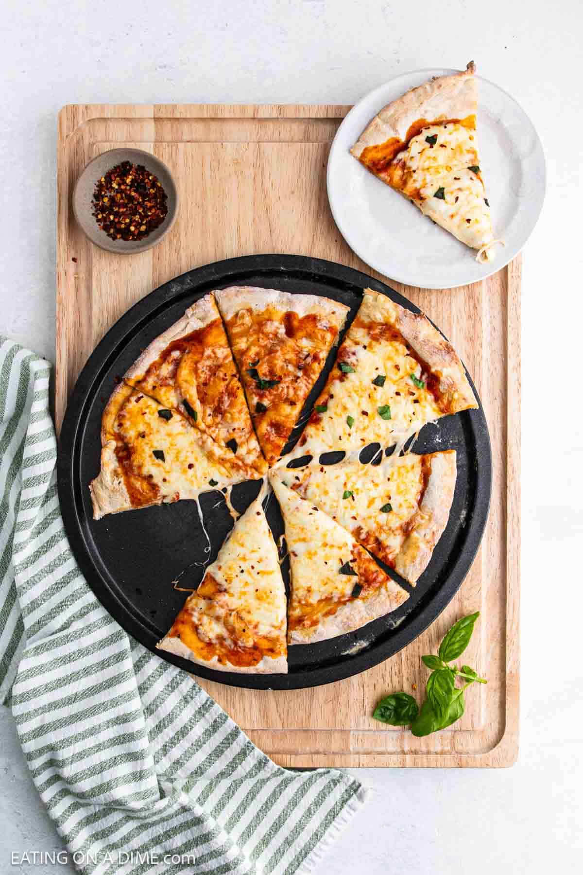 A cheese pizza with a cottage cheese pizza crust is sliced into eight pieces on a round pan atop a wooden board. One slice sits on a small white plate, surrounded by red pepper flakes, a striped towel, and fresh basil leaves.