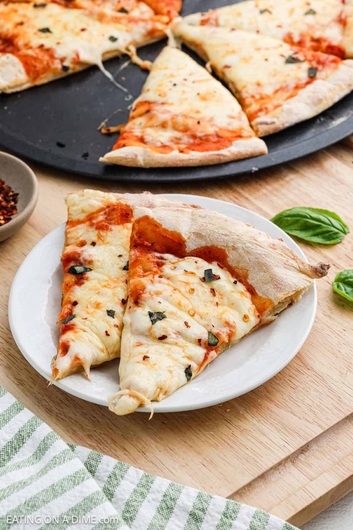 Two slices of cheese pizza with tomato sauce and basil sit on a white plate, featuring a golden pizza crust. More pizza slices rest on a pan in the background, with fresh basil leaves and a striped cloth nearby.