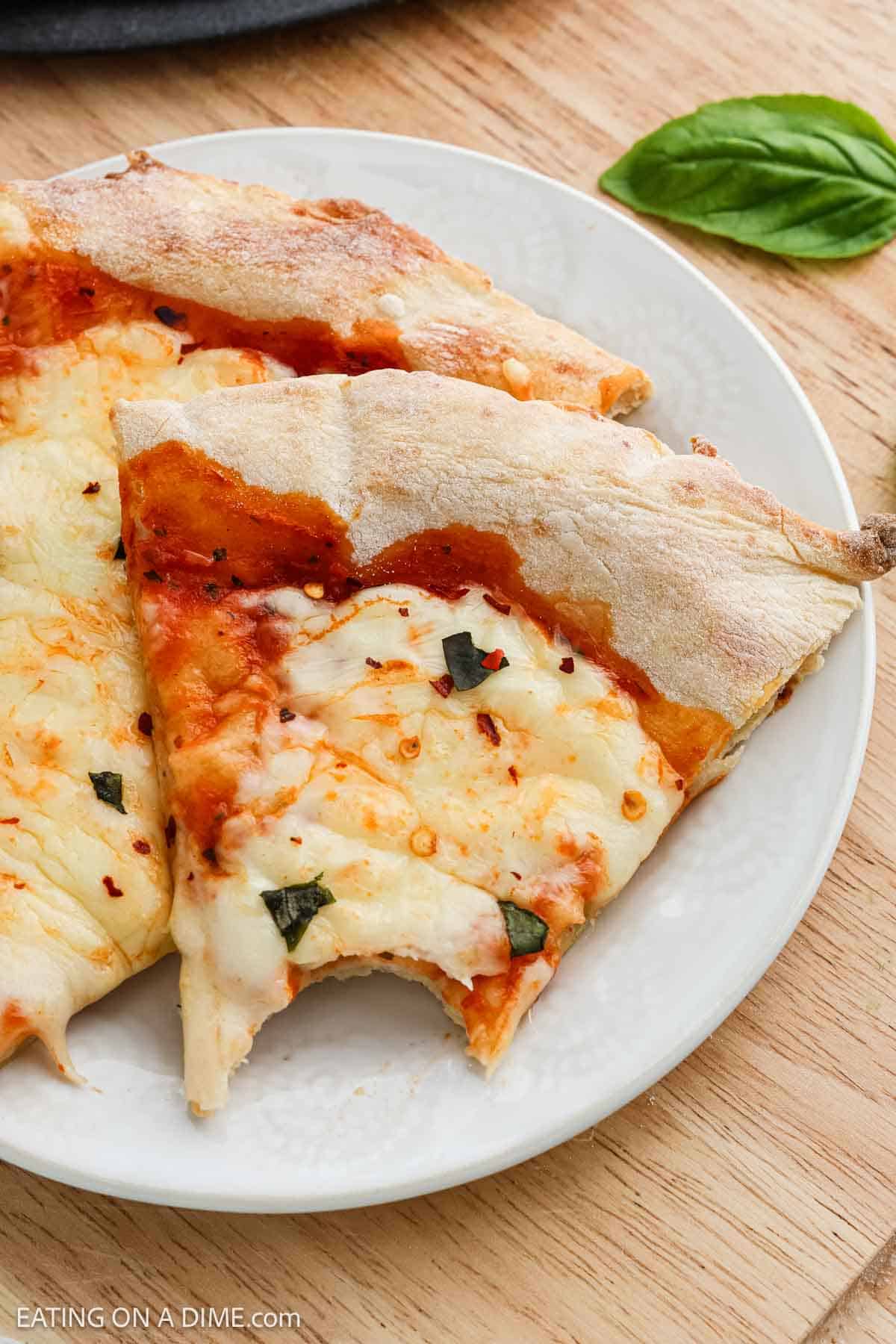 Two slices of margherita pizza with melted cheese, tomato sauce, and basil on a white plate, one slice with a bite taken out, showcase their golden pizza crust atop a wooden table.