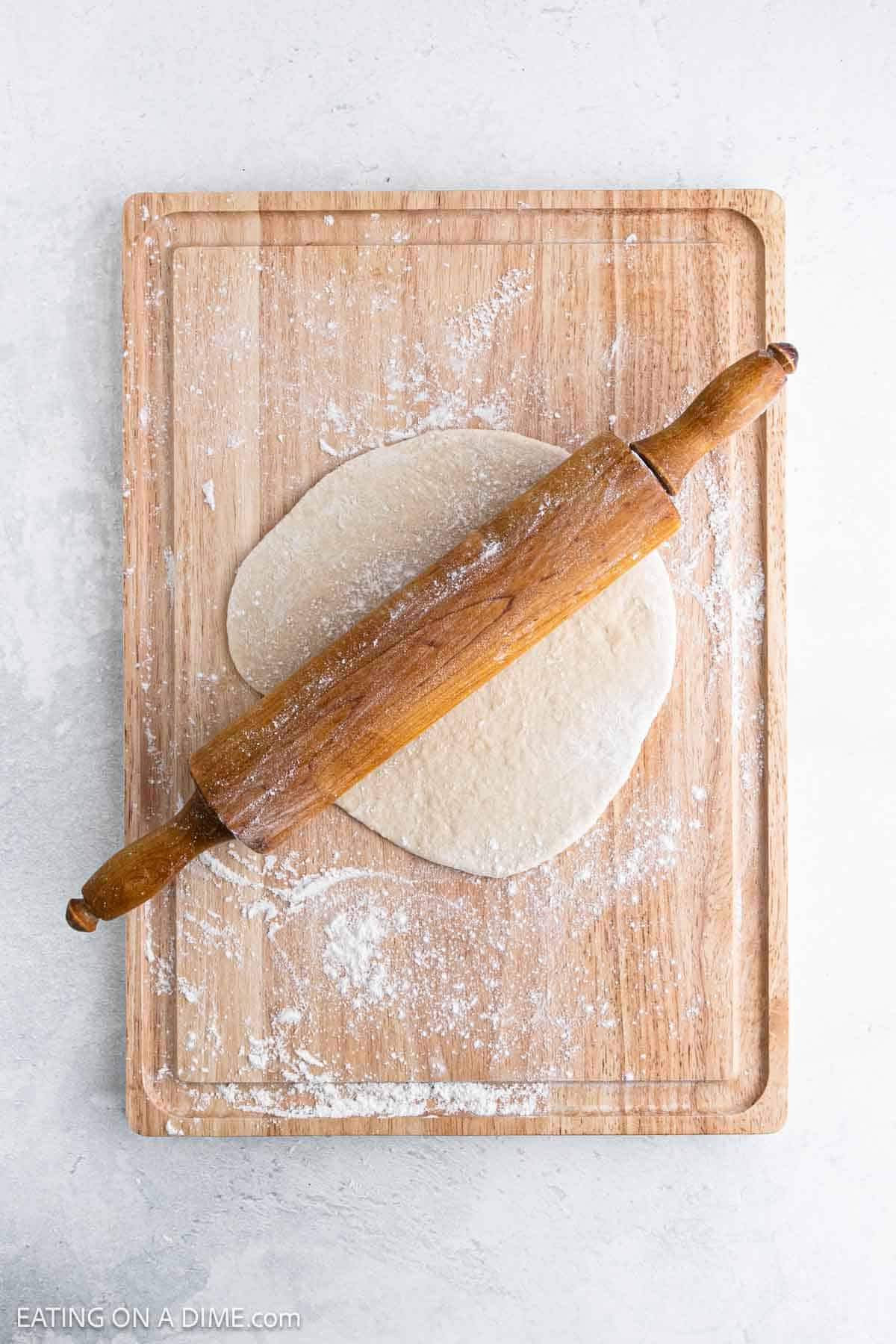A wooden rolling pin rests on top of dough for a high protein pizza crust, being rolled out on a lightly floured wooden cutting board. The surface around the board is light-colored.