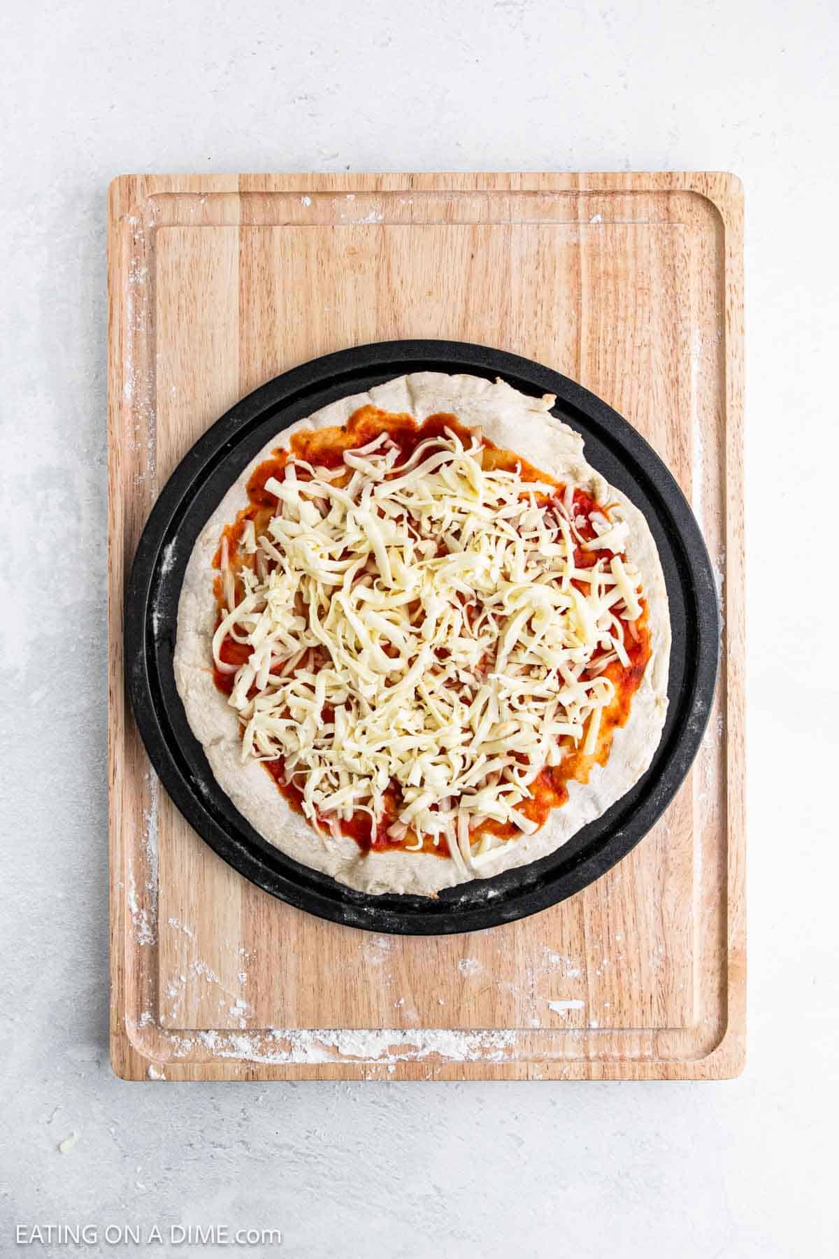 A round, uncooked pizza with tomato sauce and shredded cheese sits on a black pan atop a wooden cutting board, featuring a high protein pizza crust for a nutritious twist, against a light-colored background.