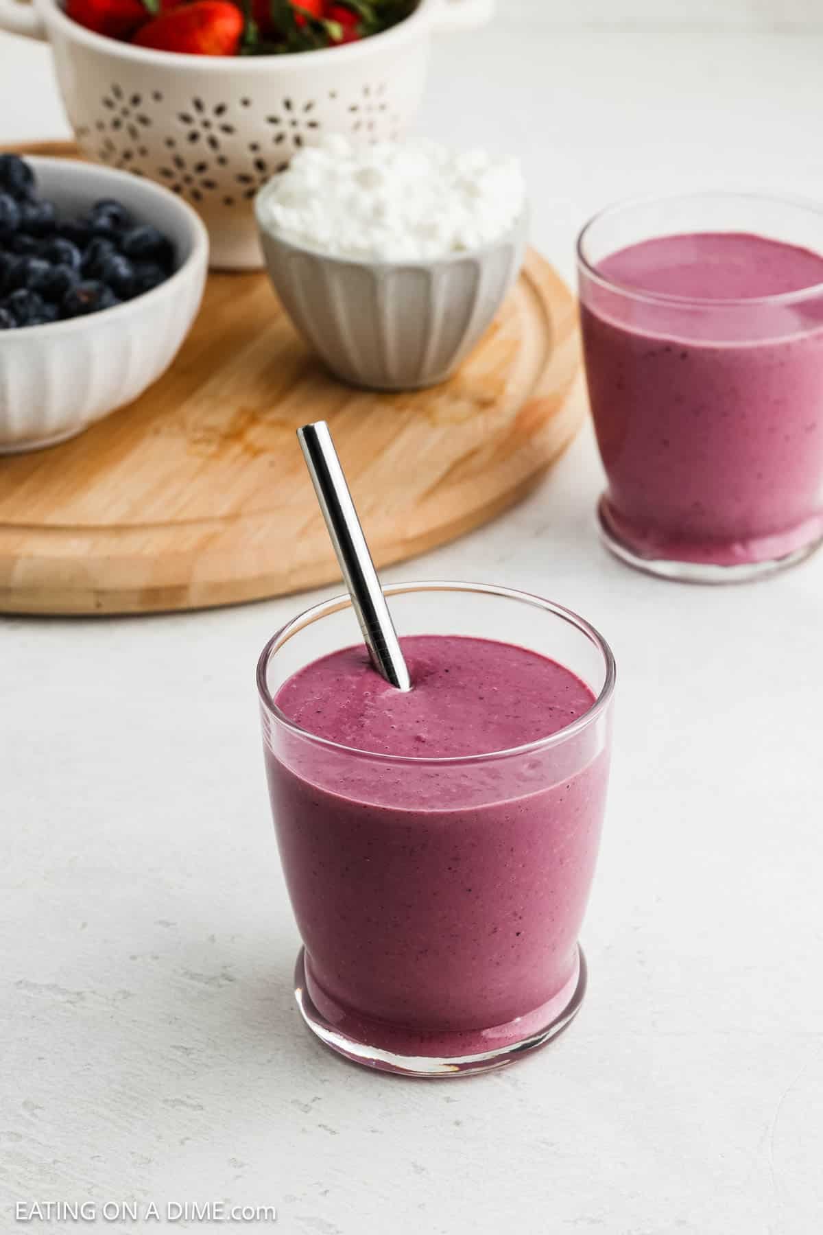 Two glasses of thick purple cottage cheese smoothie, one with a metal straw, sit on a white surface. Behind them are bowls of blueberries, strawberries, and cottage cheese on a wooden tray.