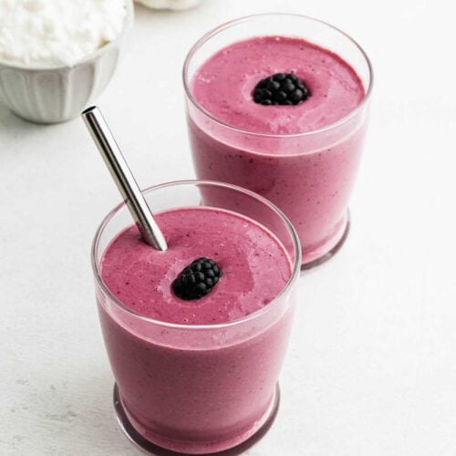 Two glasses filled with thick, pinkish-purple cottage cheese smoothies, each topped with a blackberry; one glass has a metal straw. A bowl of cottage cheese is partially visible in the background.