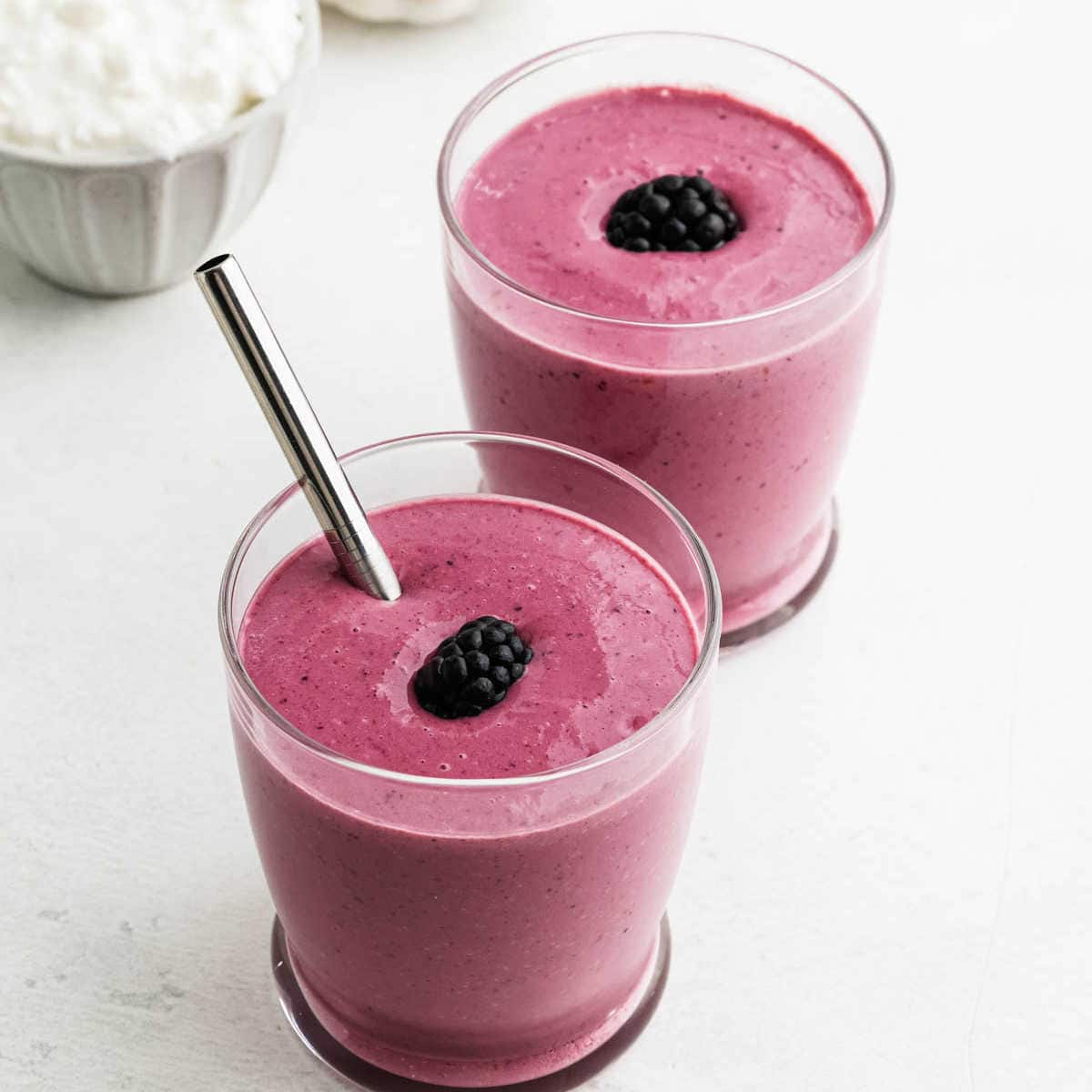 Two glasses filled with thick, pinkish-purple cottage cheese smoothies, each topped with a blackberry; one glass has a metal straw. A bowl of cottage cheese is partially visible in the background.