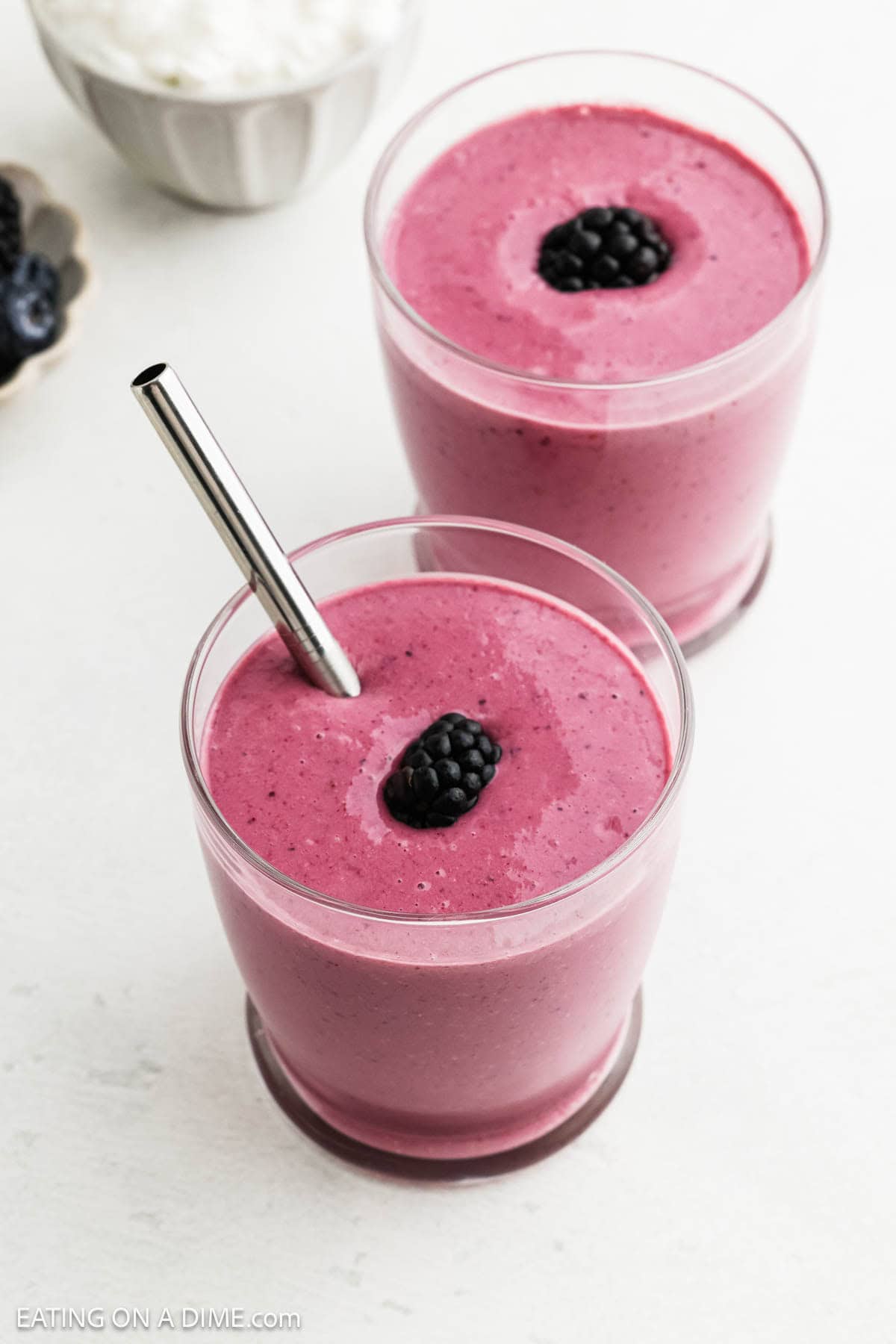 Two glasses filled with thick, pink-purple cottage cheese smoothies, each topped with a blackberry. One glass has a metal straw. The light background features a bowl of berries and a white bowl slightly visible.