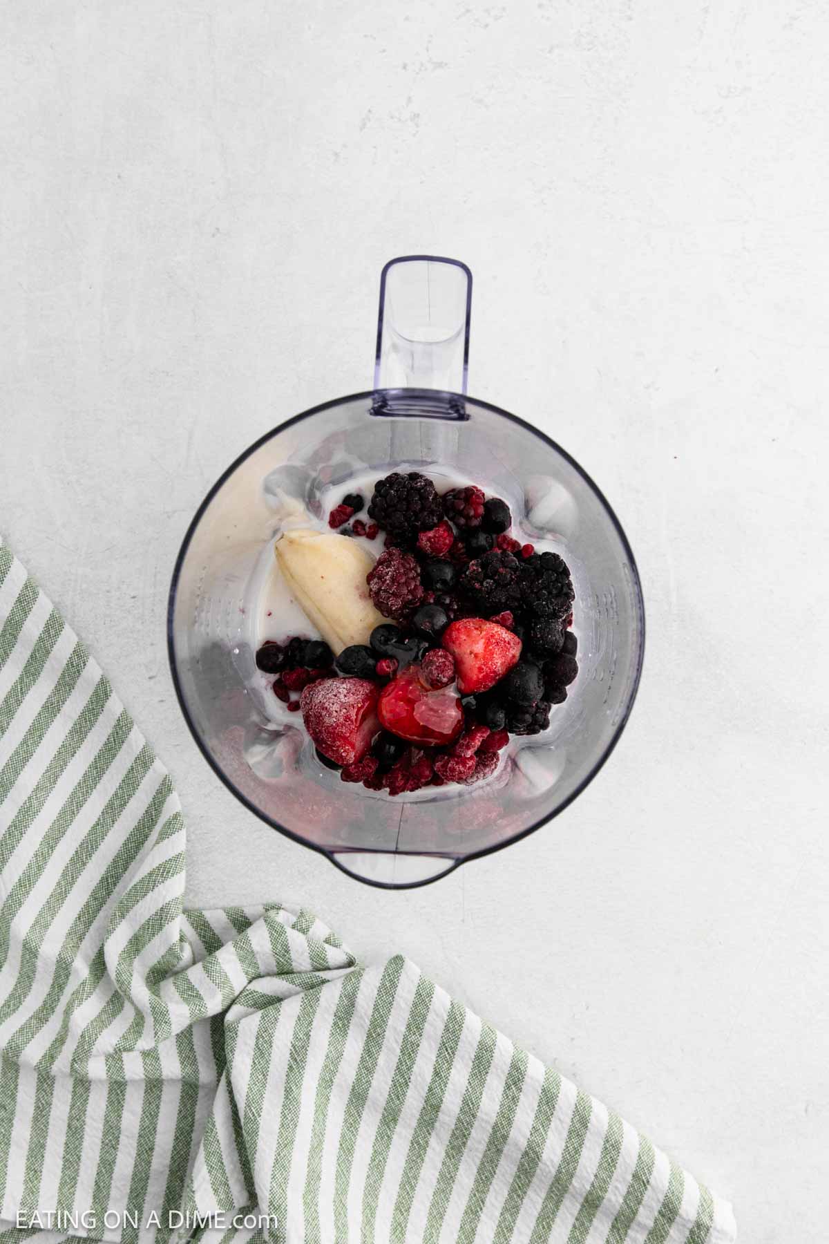 A blender filled with banana, mixed frozen berries, milk, and cottage cheese for a creamy Cottage Cheese Smoothie, viewed from above. A green and white striped cloth is partially visible on the left side.