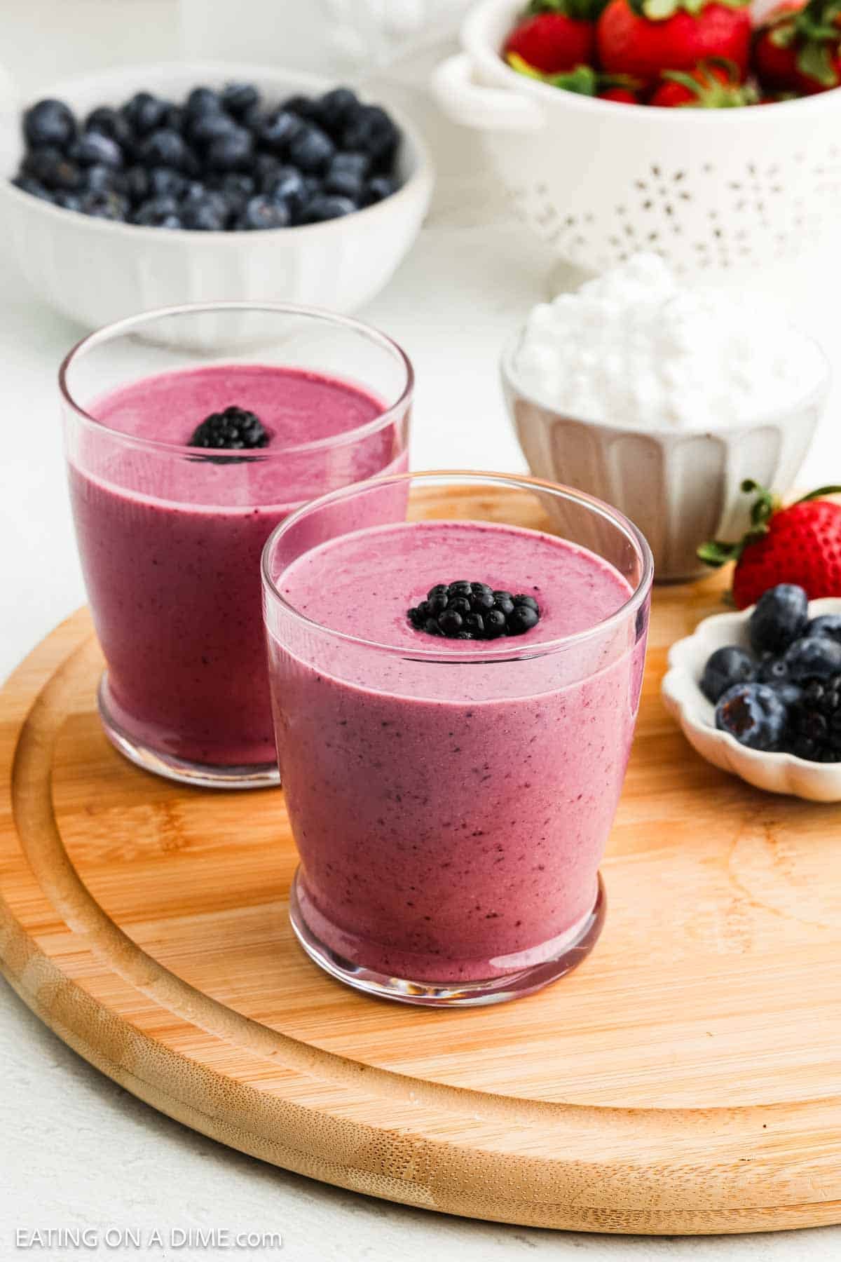 Two glasses of vibrant purple cottage cheese smoothie, each topped with a single blackberry, sit on a wooden board. In the background are bowls filled with blueberries, strawberries, and creamy cottage cheese.