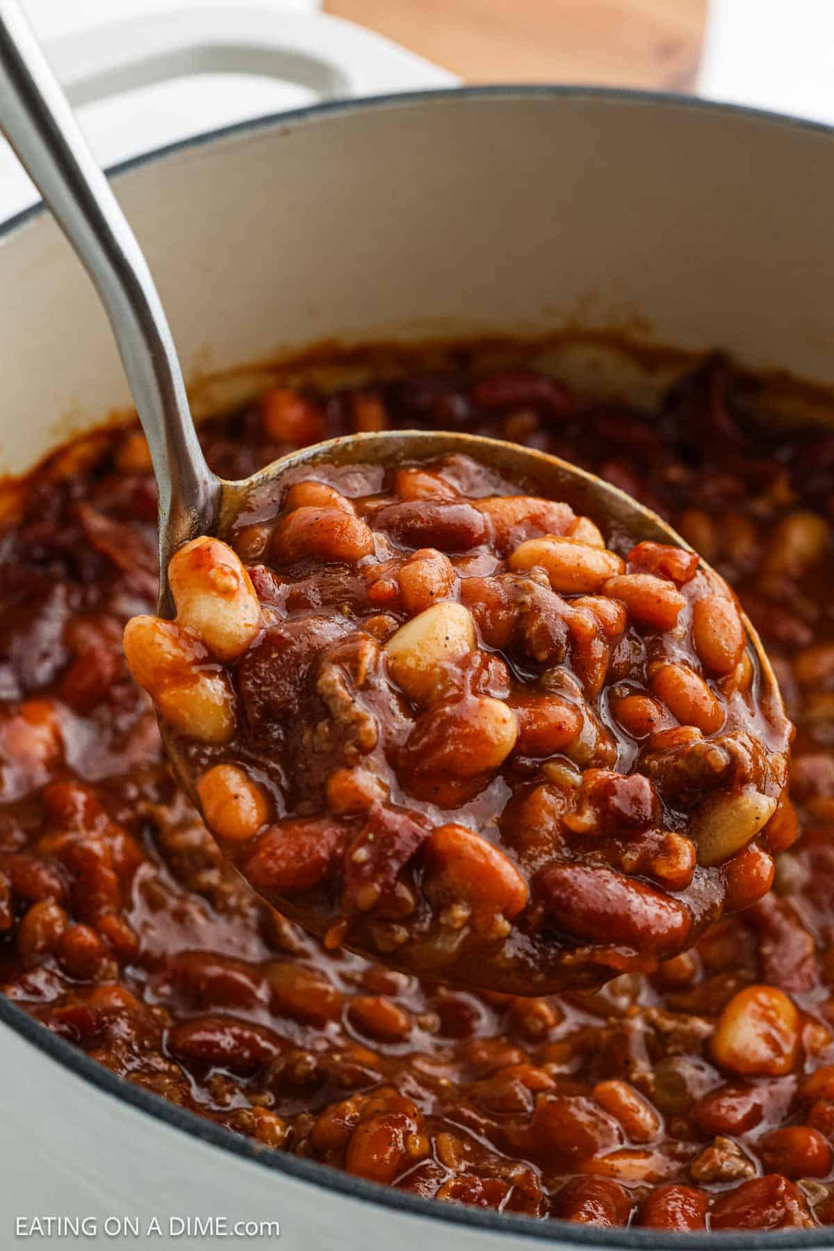 A metal ladle holds a hearty serving of Cowboy Beans in a thick, rich sauce above a white pot brimming with this classic cowboy bean recipe.