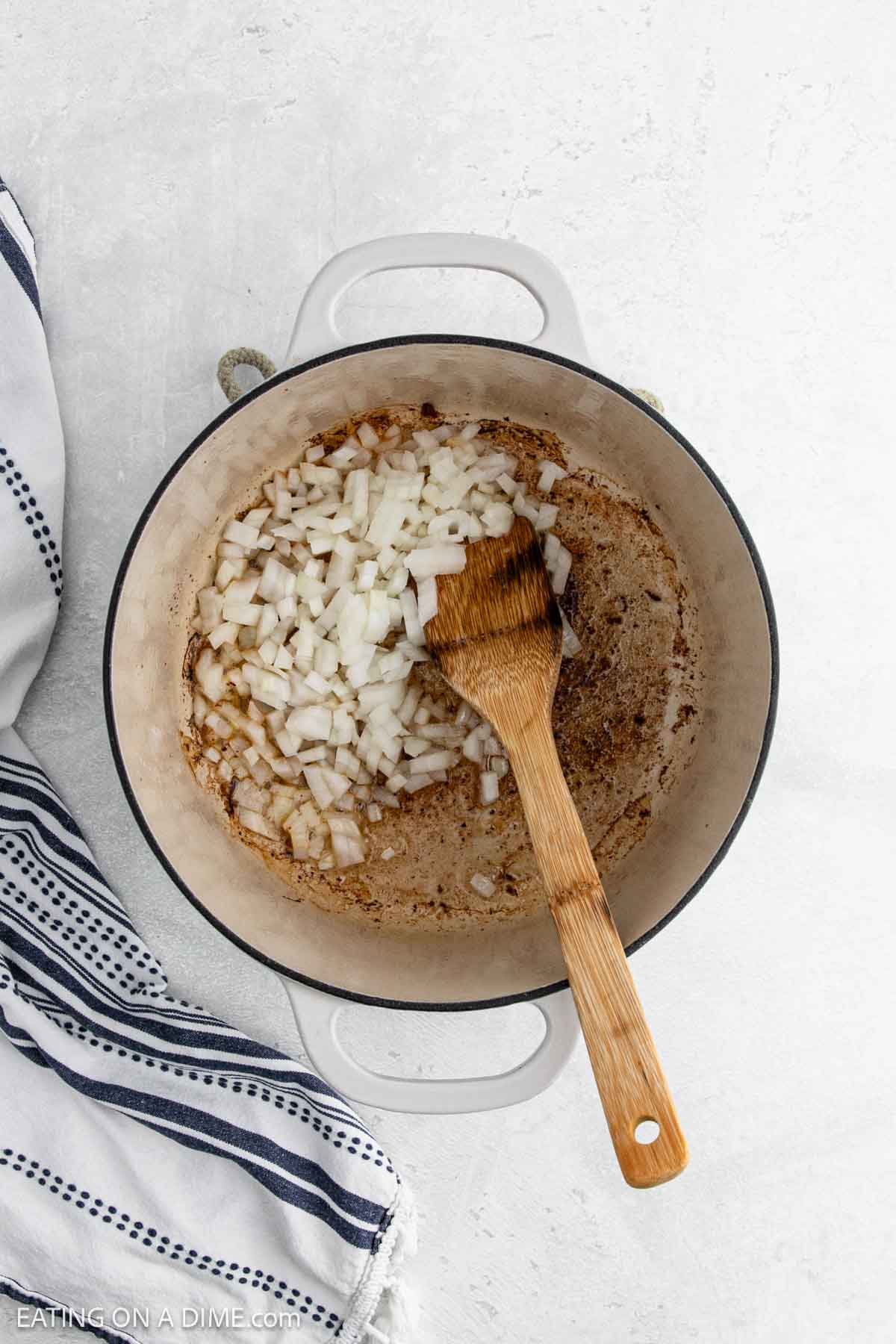 Chopped onions being sautéed in a white pot with a wooden spoon, next to a striped kitchen towel—an essential first step when preparing a hearty Cowboy Beans recipe on a light-colored surface.