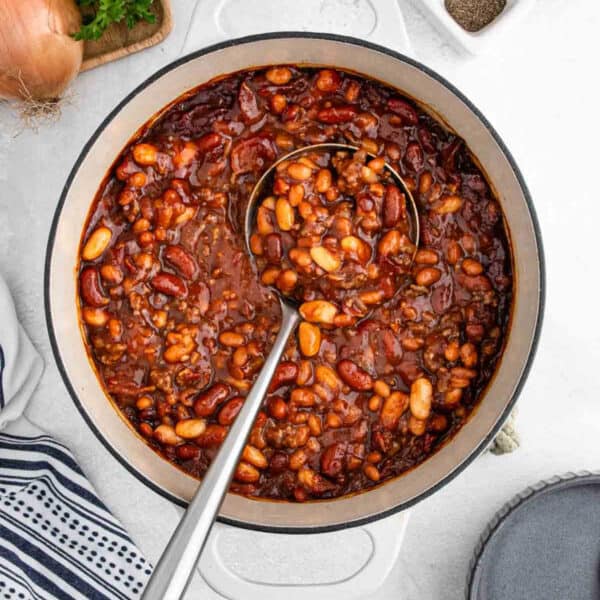 A pot of hearty cowboy food with kidney beans, white beans, ground meat, and tomato sauce sits on a white surface, ladle inside. A striped towel and onion are visible nearby—classic Cowboy Beans for any bean recipe lover.