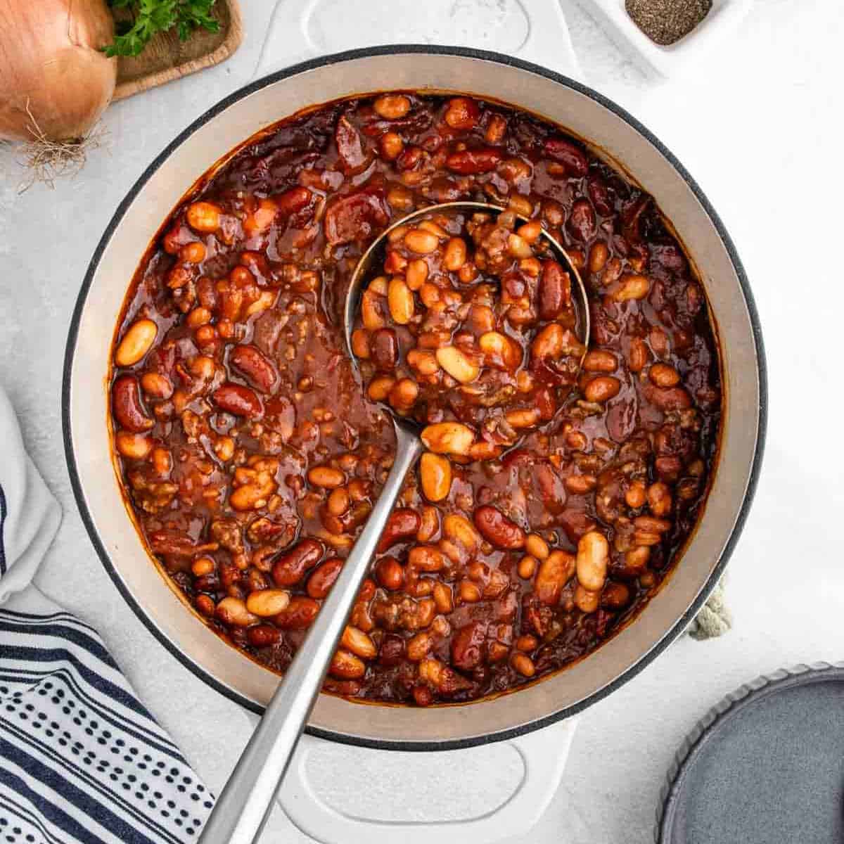 A pot of hearty cowboy food with kidney beans, white beans, ground meat, and tomato sauce sits on a white surface, ladle inside. A striped towel and onion are visible nearby—classic Cowboy Beans for any bean recipe lover.