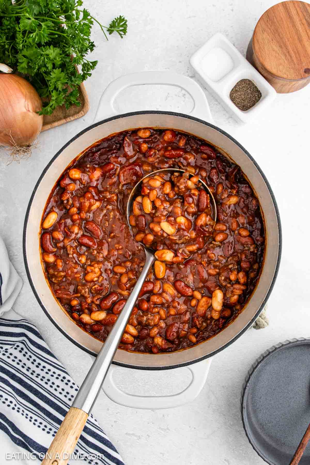 A pot of Cowboy Beans in a rich sauce with a ladle, surrounded by an onion, parsley, salt and pepper shakers, a striped towel, and an empty plate—perfect for those craving classic Western recipes.
