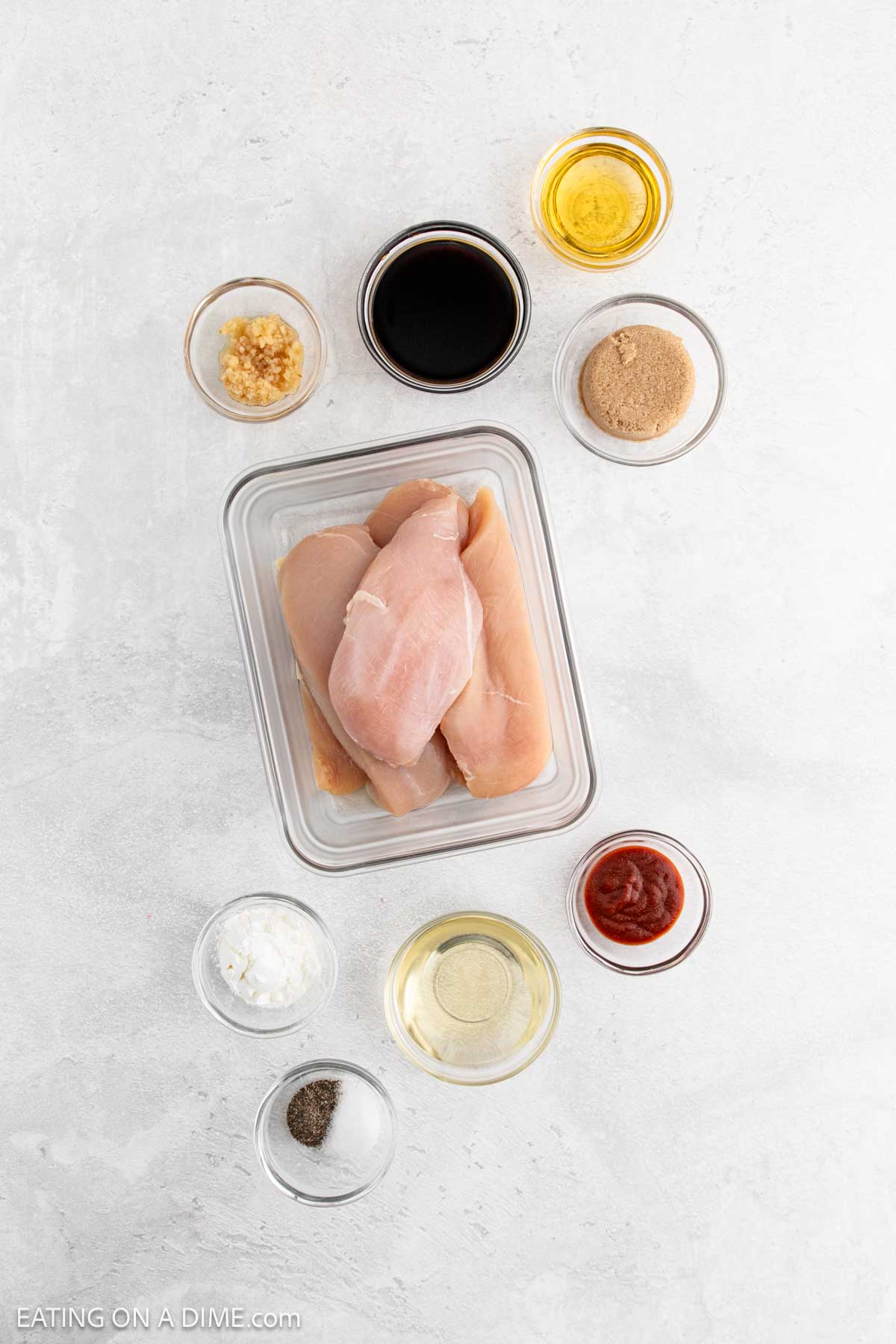 Overhead view of raw chicken breasts in a glass container, surrounded by small bowls of soy sauce, brown sugar, ketchup, minced garlic, and more—perfect ingredients for making tasty Korean BBQ Chicken on a white surface.