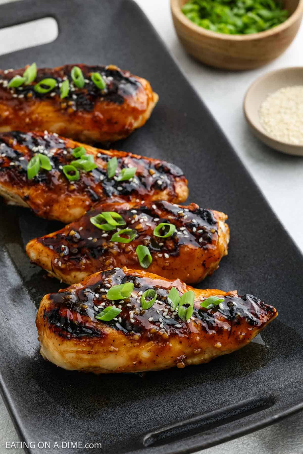 Four glazed Korean BBQ chicken breasts garnished with chopped green onions and sesame seeds are arranged on a black rectangular plate. Small bowls of chopped green onions and sesame seeds are in the background.