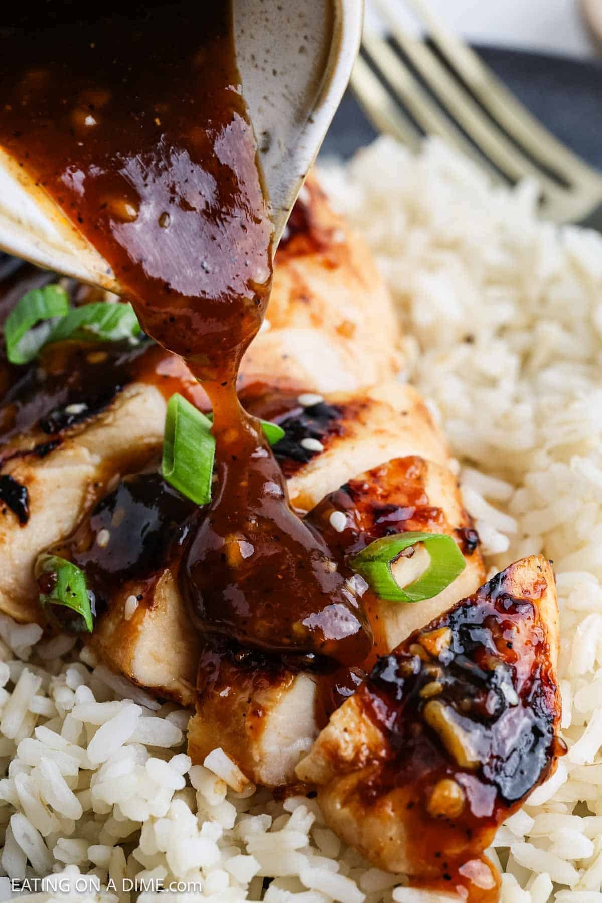 A close-up of sliced Korean BBQ chicken on a bed of white rice, being topped with a thick, glossy brown sauce and garnished with chopped green onions.