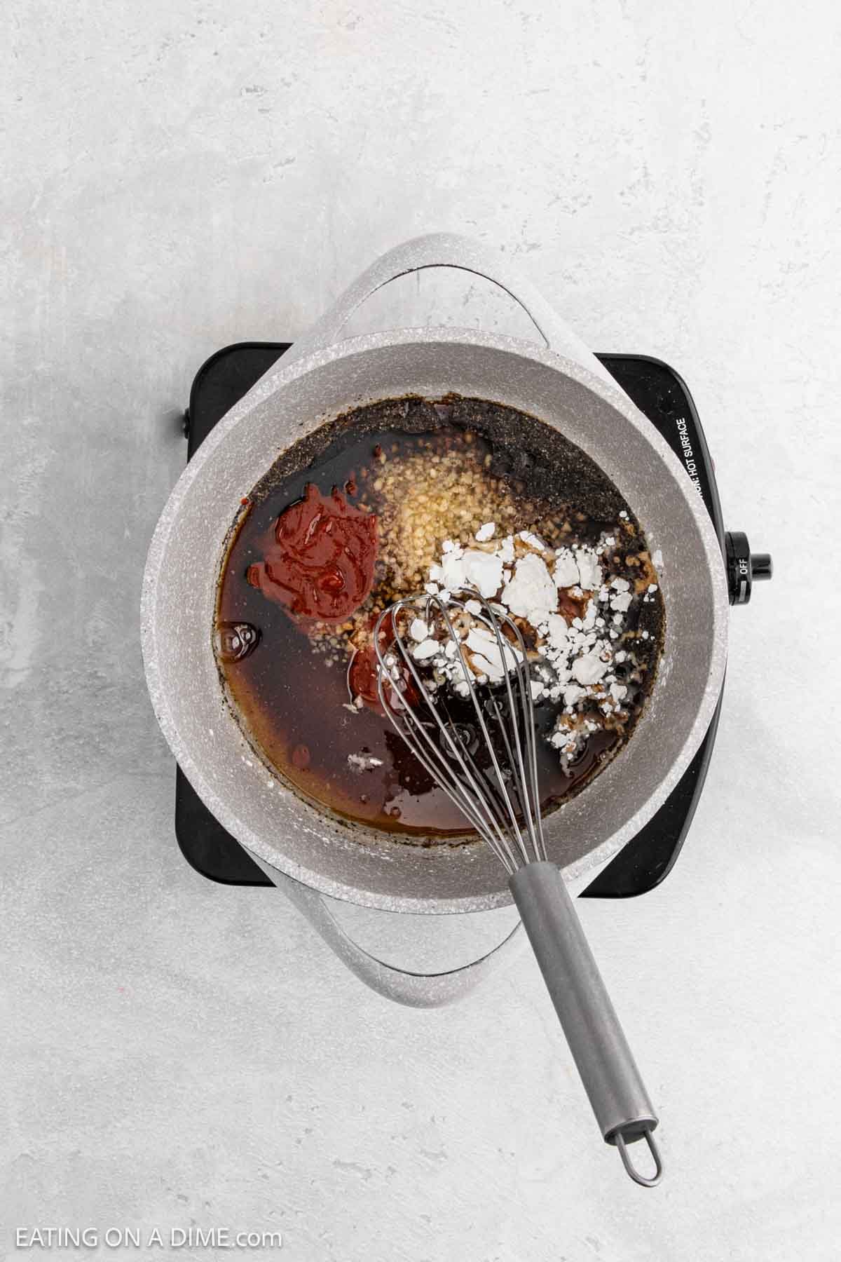 A saucepan on a hot plate contains dark sauce ingredients being mixed with a metal whisk. The sauce, perfect for Korean BBQ Chicken, includes minced garlic, brown sugar, and seasonings on a light gray background.