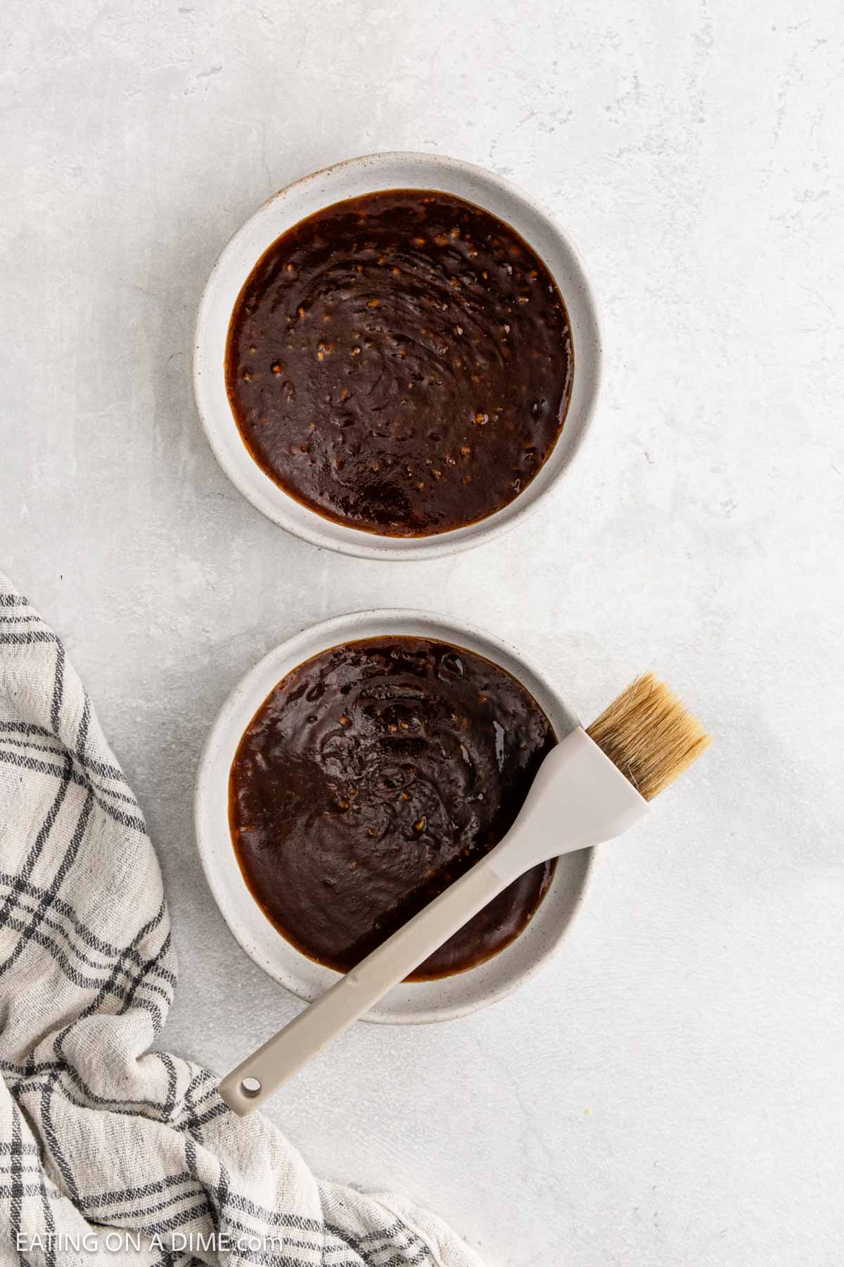 Two small bowls filled with a dark brown Korean BBQ sauce, one with a basting brush resting on top. A gray-and-white checkered cloth is partially visible beside the bowls on a light textured surface.