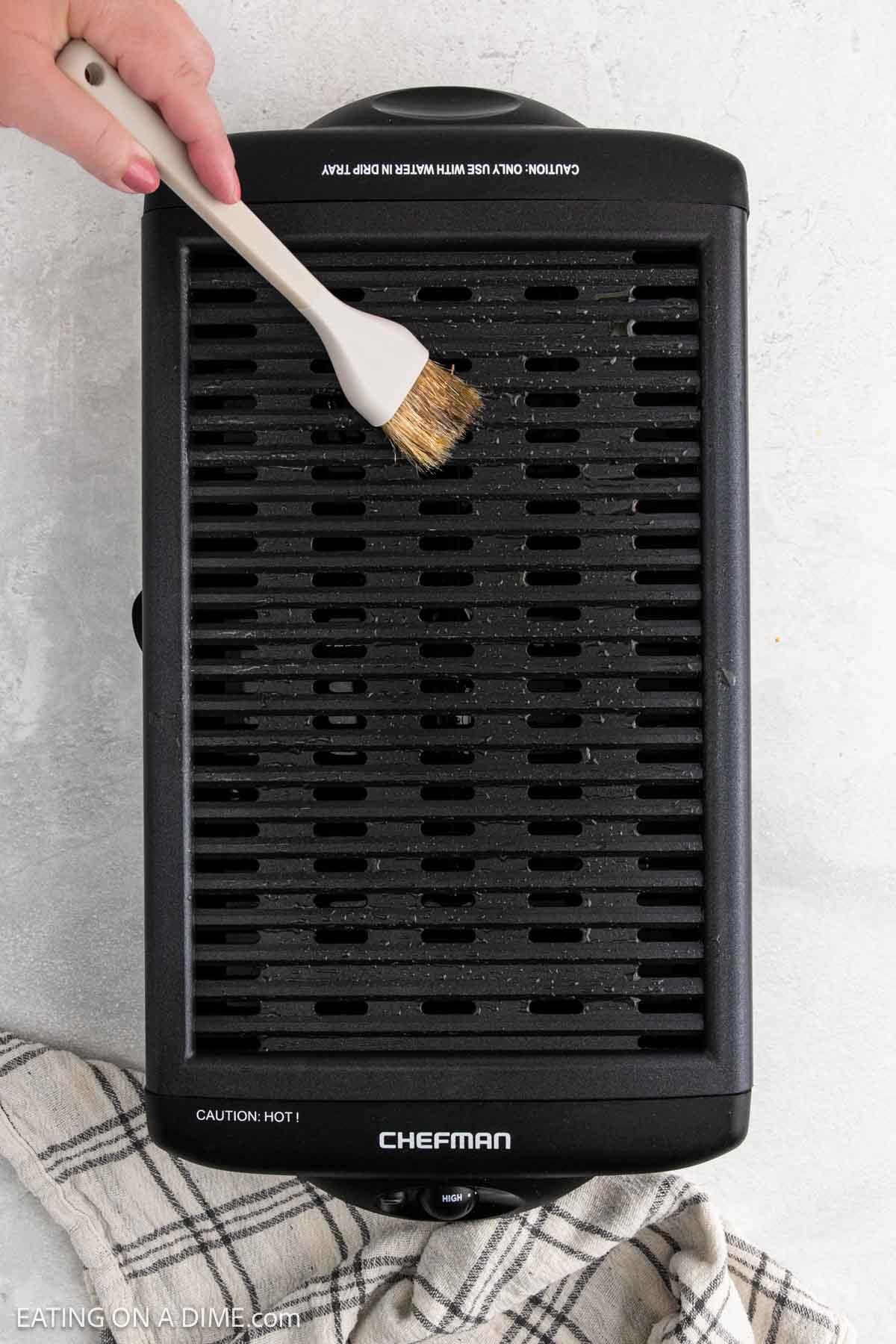 A hand uses a brush to clean an electric Chefman grill, which is placed on a light surface next to a striped kitchen towel—perfect for whipping up Korean BBQ or BBQ Chicken at home.