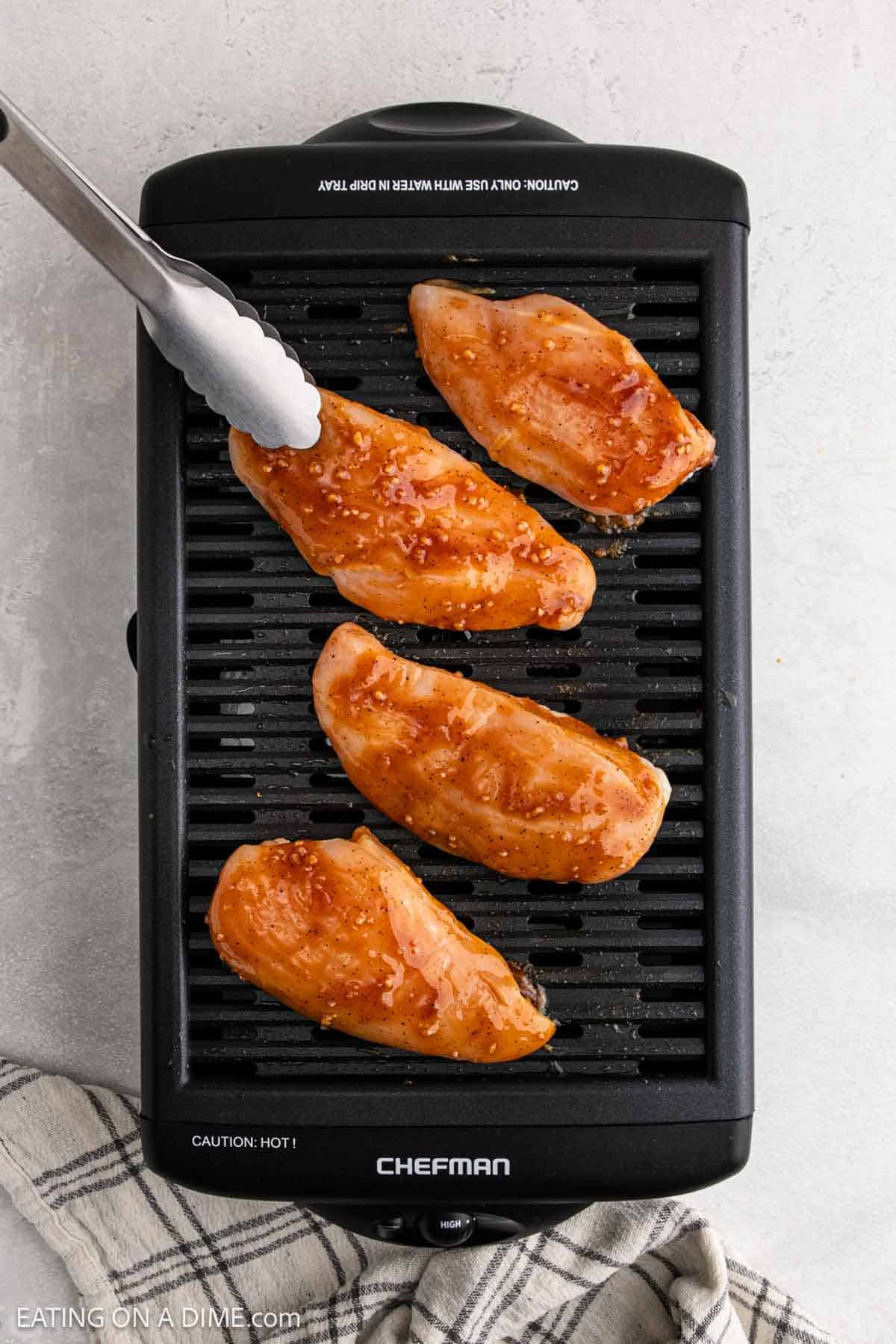 Four raw, marinated Korean BBQ chicken breasts are placed on an electric grill with metal tongs holding one piece. A checkered kitchen towel is nearby on the light-colored countertop.