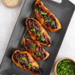 Four grilled Korean BBQ chicken breasts topped with a glossy sauce and garnished with sliced green onions are arranged on a dark rectangular platter. Bowls of sesame seeds and chopped green onions sit nearby.