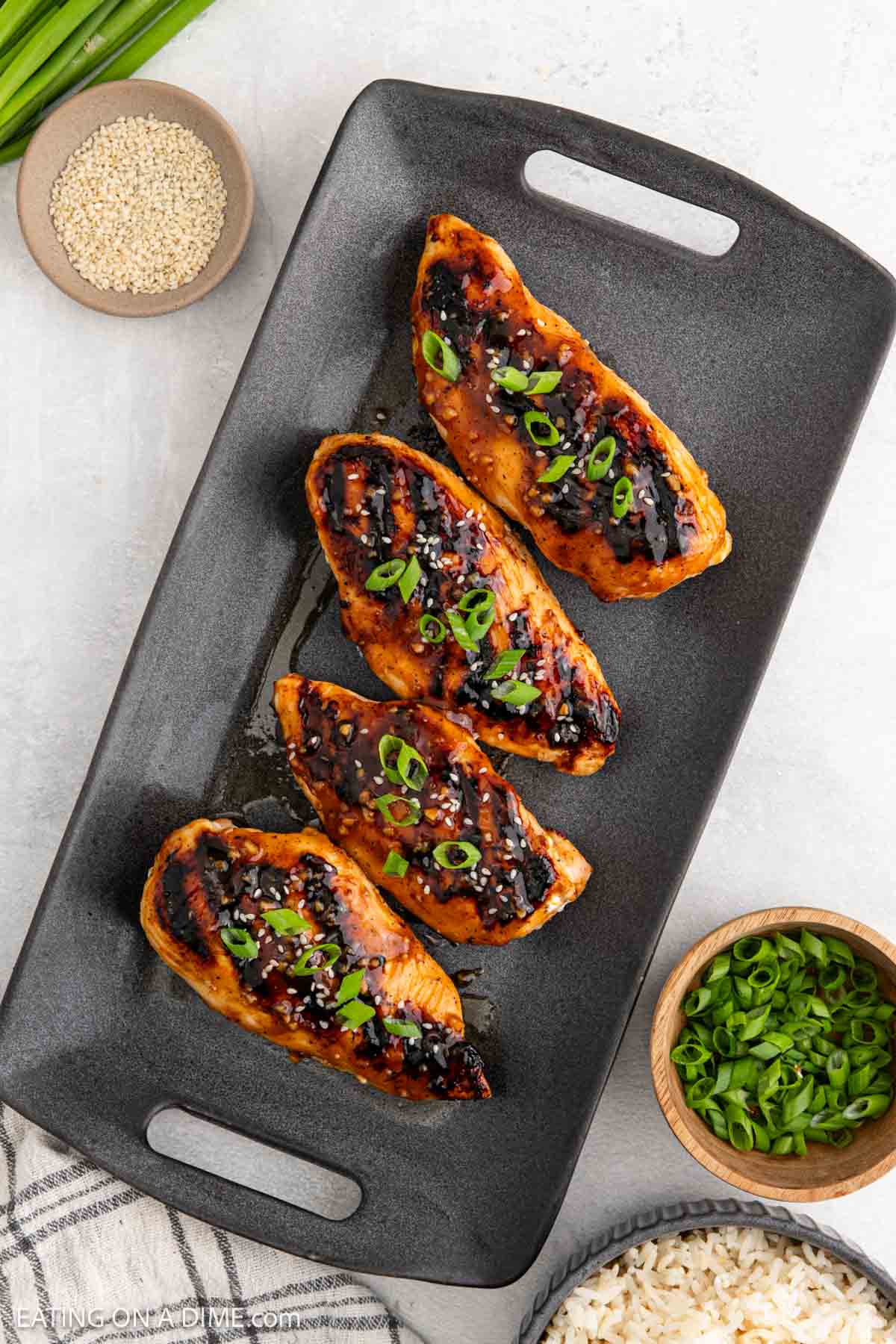 Four glazed and grilled Korean BBQ chicken breasts are arranged on a black rectangular tray, garnished with chopped green onions. Small bowls of sesame seeds, sliced green onions, and white rice are nearby.