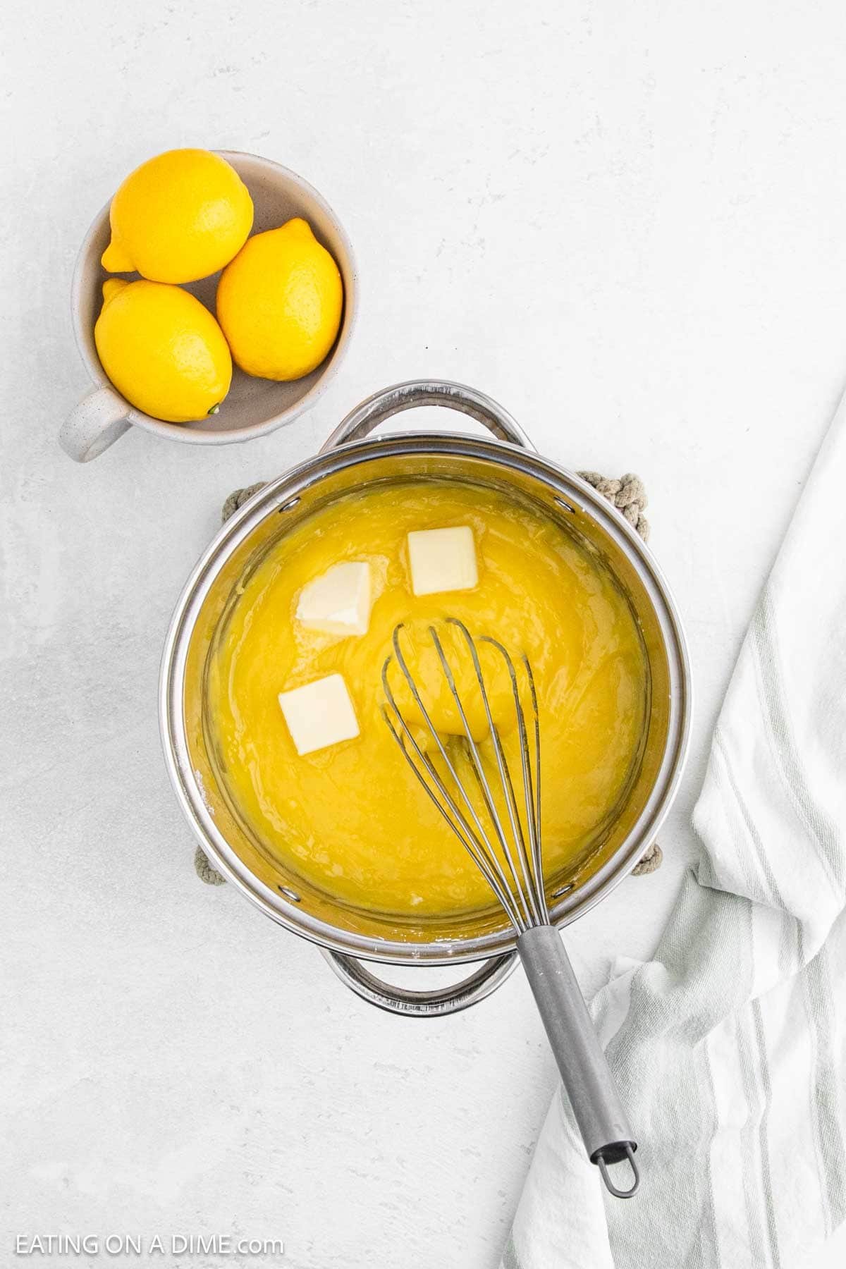 Adding slices of butter into the lemon mixture in a large pot with a whisk with a small bowl of lemons on the side