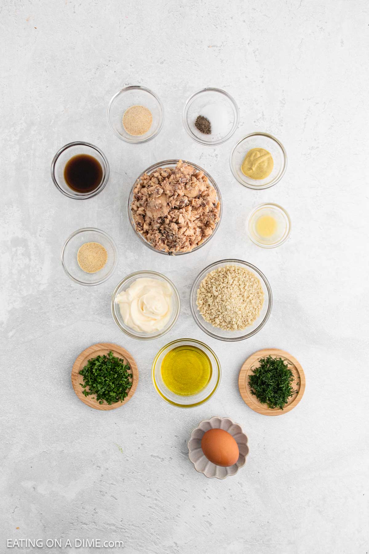 Overhead view of neatly arranged ingredients for Salmon Patties in small bowls on a white surface, including canned tuna, mayonnaise, breadcrumbs, an egg, chopped parsley, olive oil, spices, and seasonings.