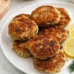 A white plate with seven golden-brown salmon patties arranged in a cluster, garnished with three lemon slices on the side—perfect for fans of easy salmon recipes.