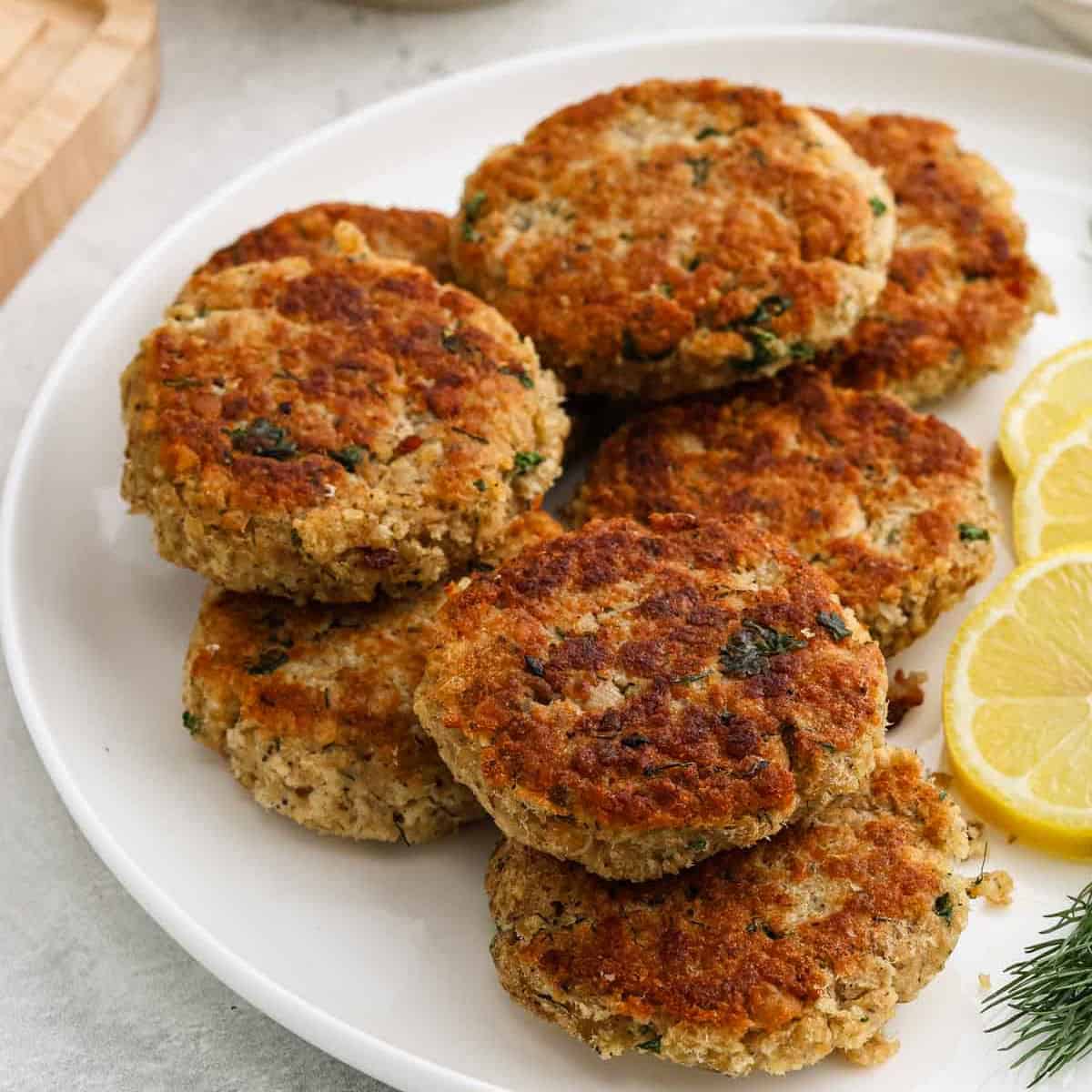 A white plate with seven golden-brown salmon patties arranged in a cluster, garnished with three lemon slices on the side—perfect for fans of easy salmon recipes.