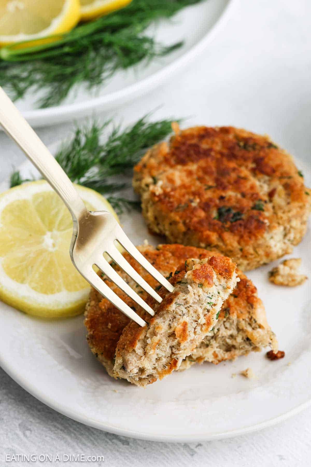 A fork holds a piece of golden-brown salmon patty on a white plate, accompanied by lemon slices and fresh dill, with another whole salmon cake in the background—perfect for fans of classic salmon recipes.
