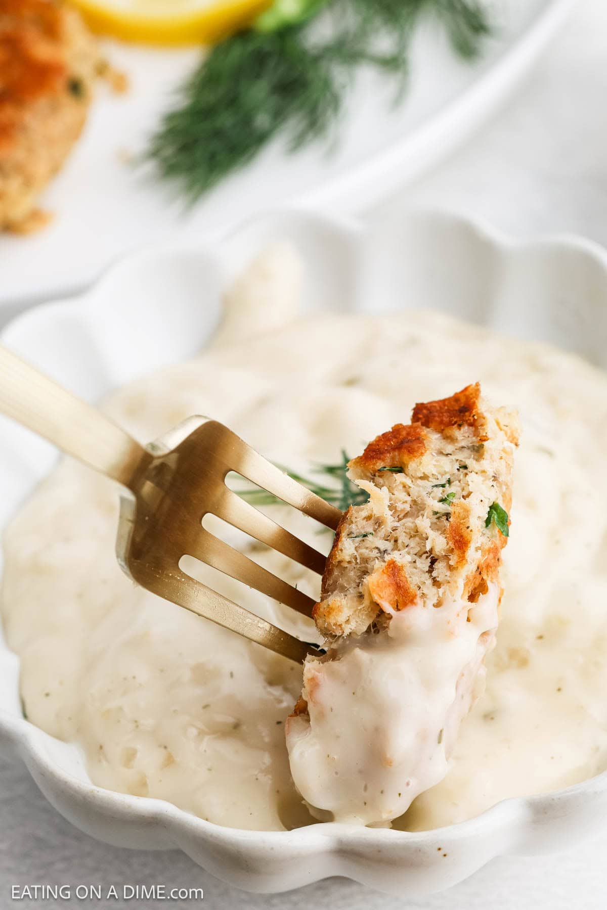 A fork holds a bite of golden-brown salmon patties dipped in creamy white sauce, with more sauce in a white bowl below. Fresh dill and lemon wedges are blurred in the background.