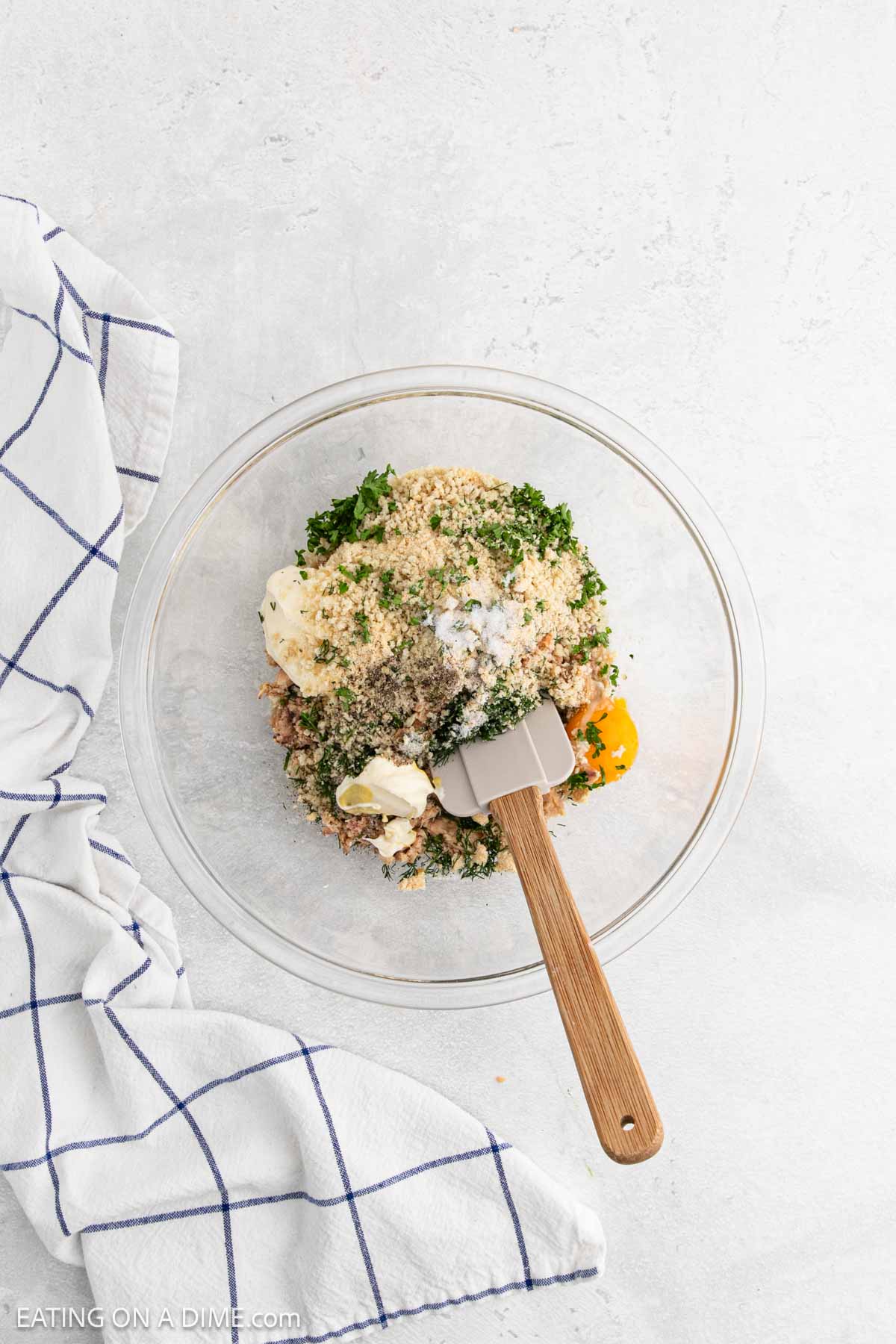 A glass bowl containing breadcrumbs, herbs, mayonnaise, and eggs being mixed with a spatula—ingredients coming together for Salmon Cakes—sits next to a white kitchen towel with blue stripes on a light surface.