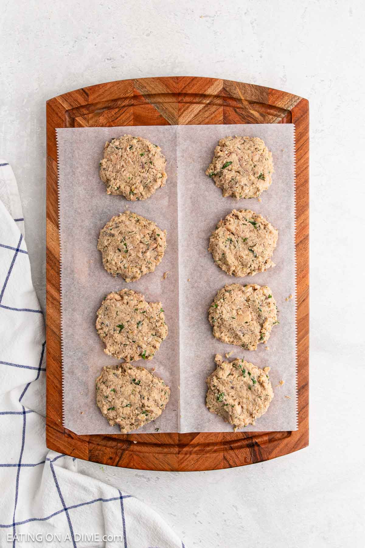 Eight uncooked tuna and salmon patties are arranged in two rows on a piece of parchment paper atop a wooden cutting board. A white kitchen towel with blue lines is partially visible on the left side.