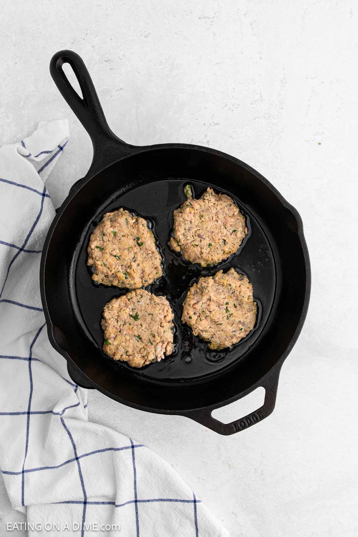 A cast iron skillet with four uncooked salmon patties sits on a white surface next to a white kitchen towel with blue stripes.