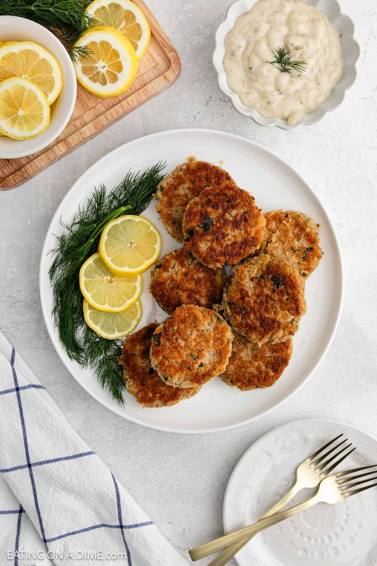 A white plate with golden-brown salmon patties, garnished with fresh dill and lemon slices. Nearby are a bowl of creamy dipping sauce, a plate with forks, and extra lemon slices—a truly easy salmon recipe for any occasion.