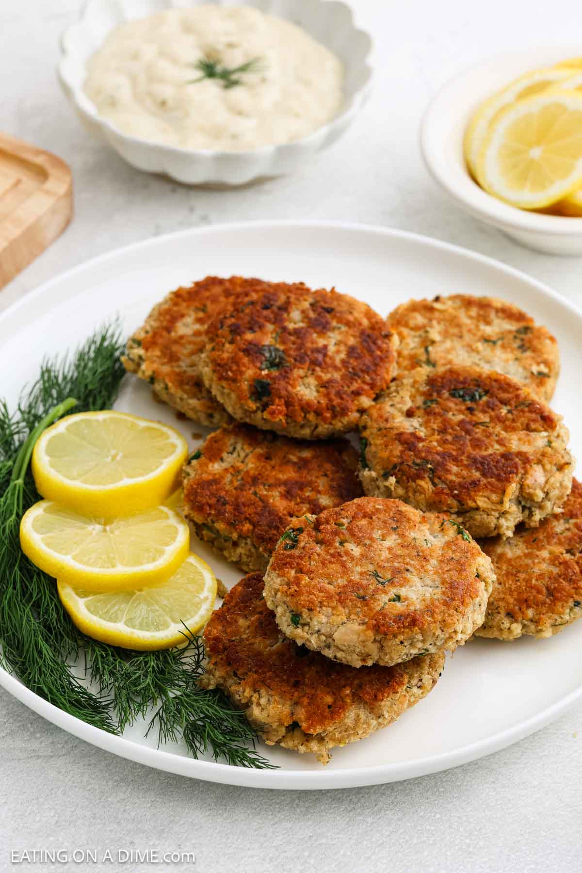 A white plate with golden-brown salmon patties, garnished with fresh dill and lemon slices. In the background, a bowl of dipping sauce sits next to a plate of lemon wedges, complementing the delicious salmon patties.
