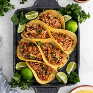 A black tray holds six corn tortillas filled with shredded slow cooker chicken tinga, garnished with chopped cilantro and surrounded by lime wedges and fresh cilantro sprigs.