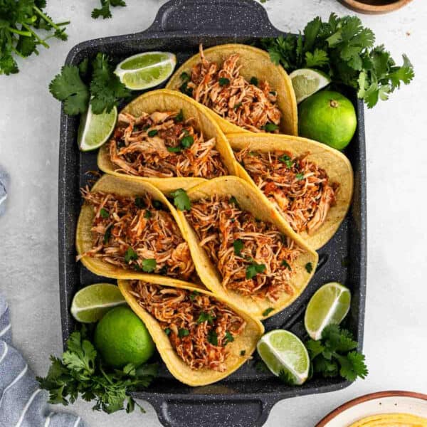 A black tray holds six corn tortillas filled with shredded slow cooker chicken tinga, garnished with chopped cilantro and surrounded by lime wedges and fresh cilantro sprigs.
