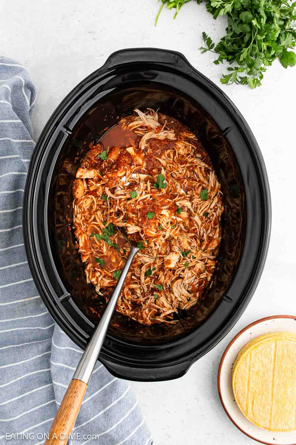 Overhead view of Slow Cooker Chicken Tinga topped with cilantro, a serving spoon, a stack of corn tortillas, fresh parsley, and a striped kitchen towel nearby.