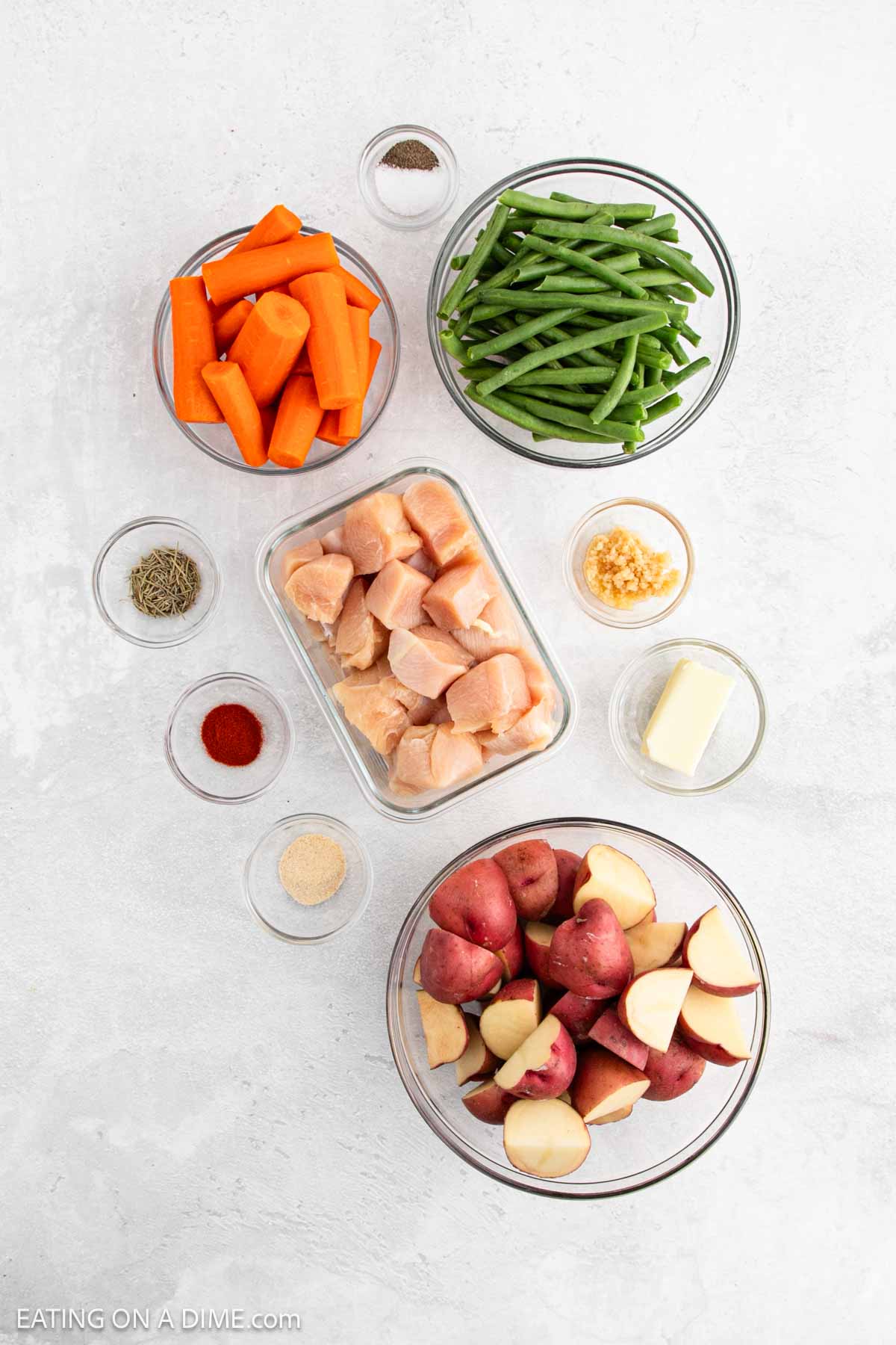 Bowls with raw cubed chicken, fresh vegetables like halved red potatoes, cut carrots, green beans, minced garlic, a pat of butter, and small bowls of spices on a white surface—perfect for a slow cooker meal.