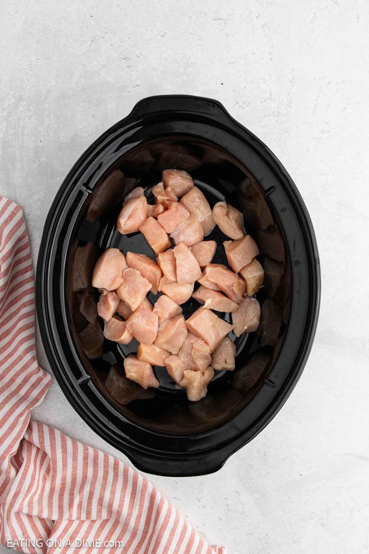 Overhead view of raw, cubed chicken pieces placed in a black slow cooker on a light surface, perfect for Slow Cooker Chicken or other Slow Cooker Recipes. A pink and white striped cloth sits to the side.