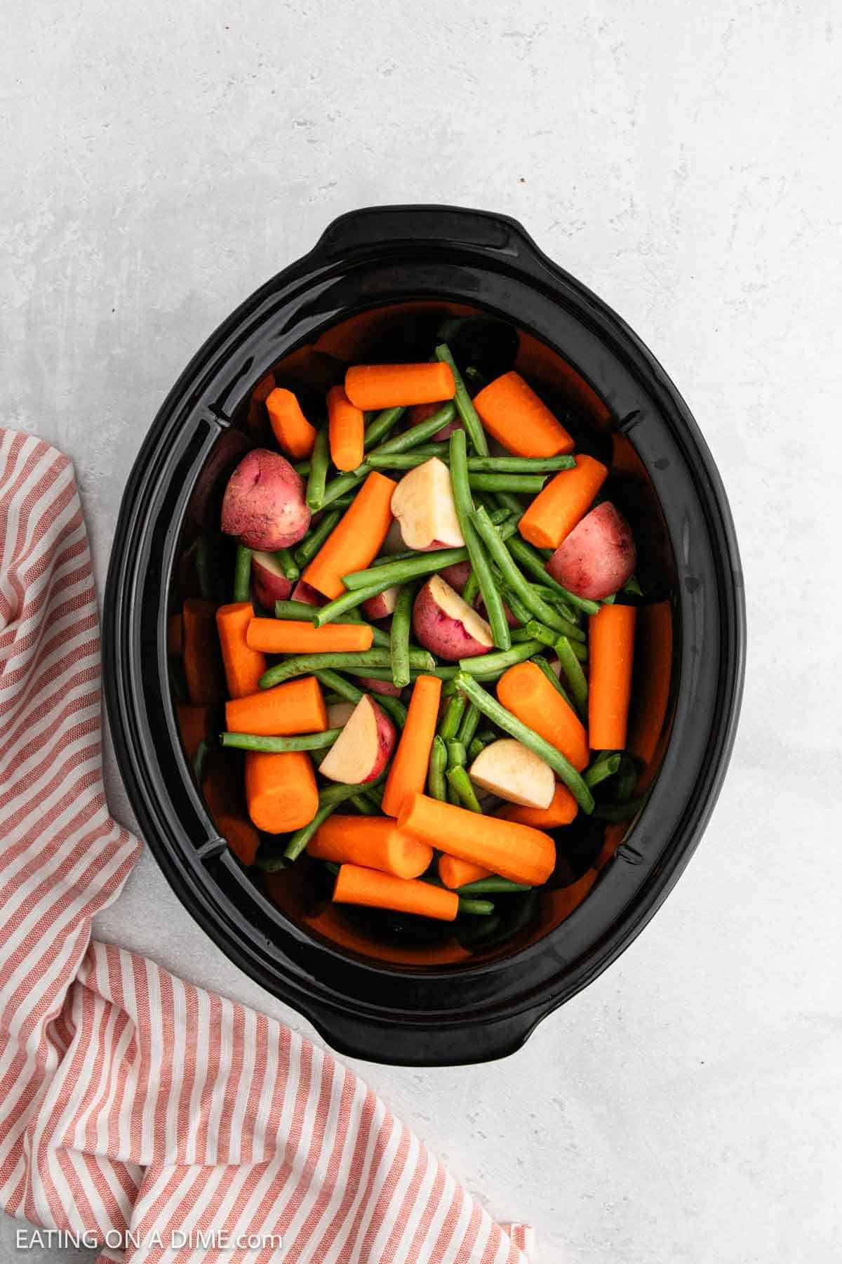 A slow cooker filled with uncooked vegetables and tender chicken, including carrots, green beans, potatoes, and garlic, sits on a light countertop beside a pink and white striped kitchen towel.