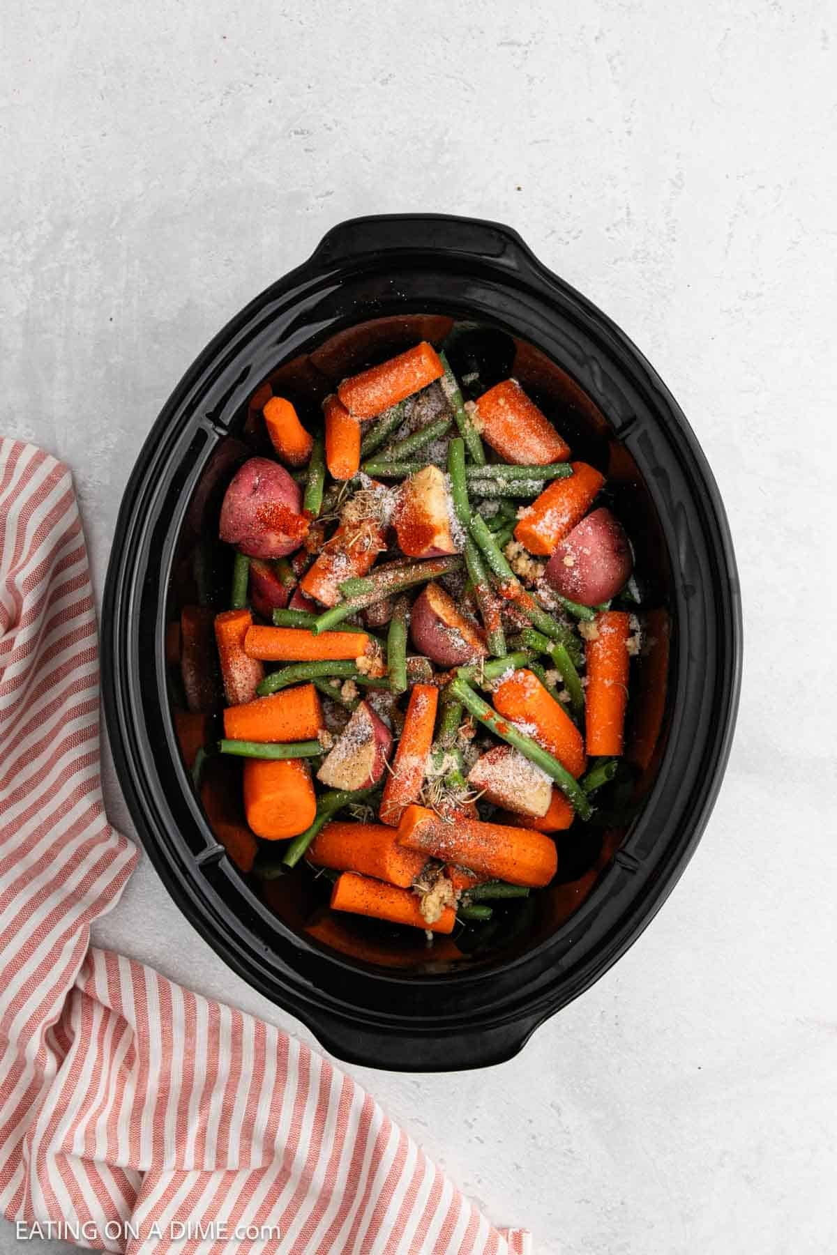 A slow cooker filled with chopped carrots, green beans, red potatoes, and seasonings—perfect for hearty Slow Cooker Chicken and Vegetables—sits on a light surface next to a pink and white striped cloth.