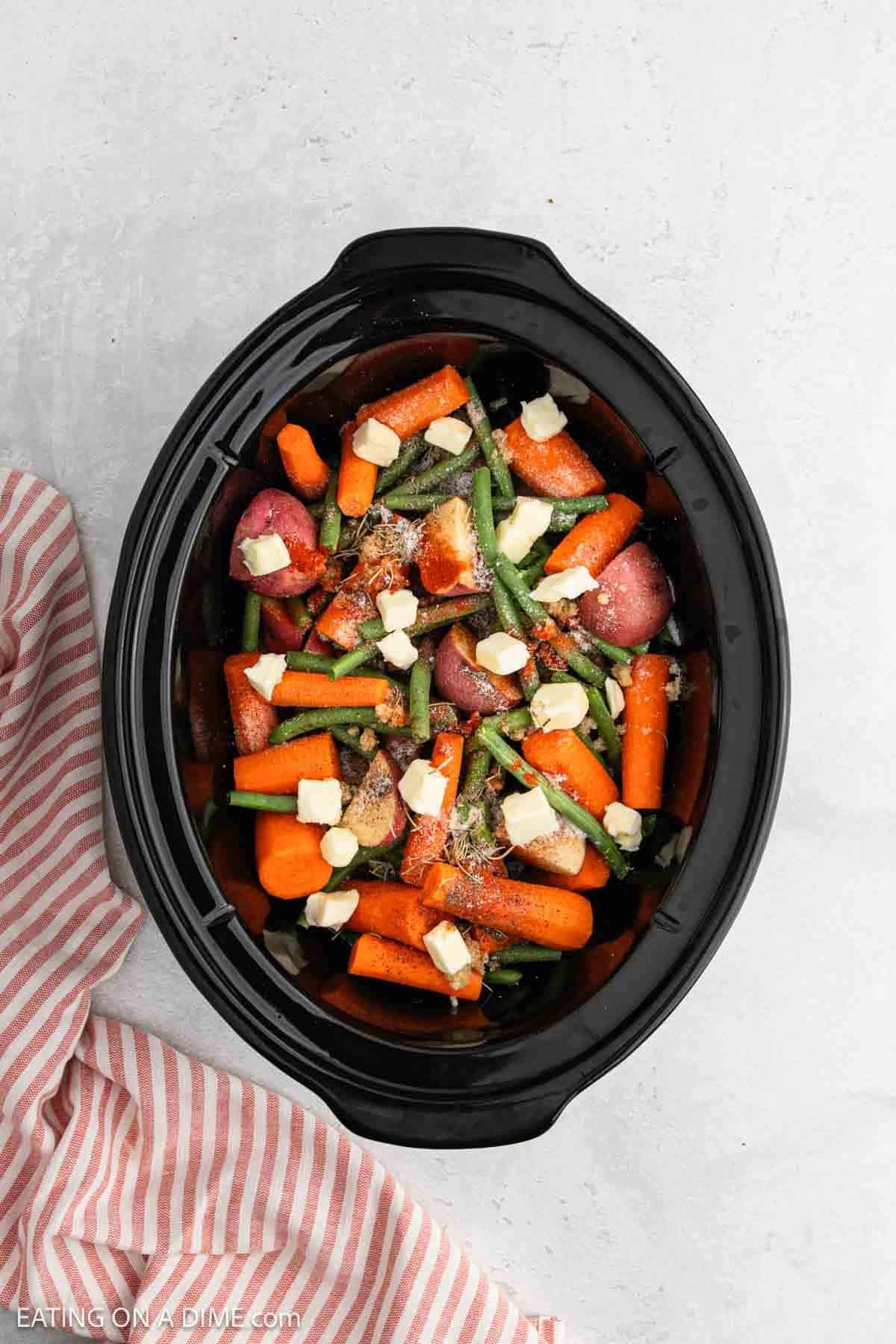 Overhead view of a slow cooker filled with raw carrots, green beans, red potatoes, and chunks of butter, ready to cook—perfect for easy slow cooker chicken and vegetables. A red and white striped cloth sits beside it on a light surface.