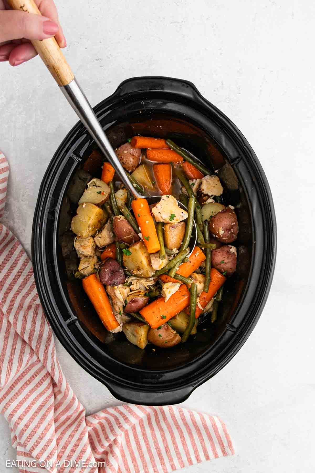 A hand uses a ladle to serve hearty Slow Cooker Chicken and Vegetables stew—filled with potatoes, carrots, and green beans—from a black slow cooker. A striped towel is partially visible on the side.