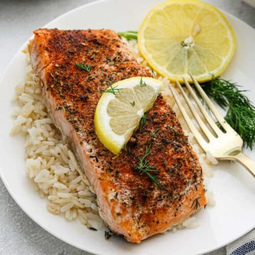 A plate with a seasoned, Slow Cooker Salmon fillet on a bed of rice, garnished with fresh dill and lemon slices, with a gold fork on the side.