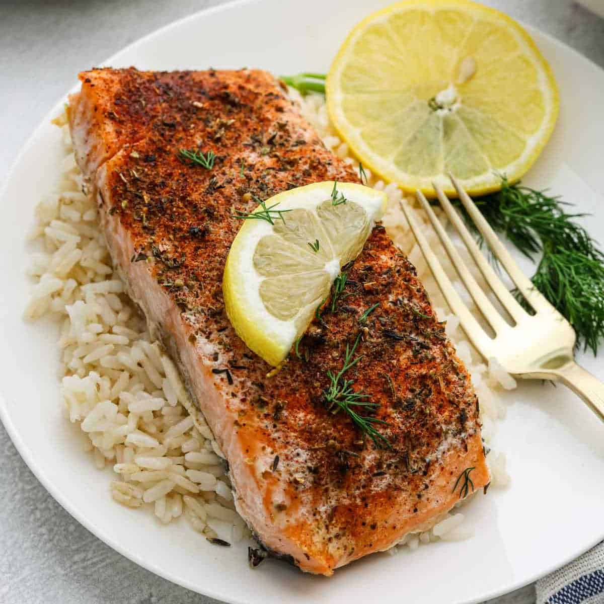 A plate with a seasoned, Slow Cooker Salmon fillet on a bed of rice, garnished with fresh dill and lemon slices, with a gold fork on the side.